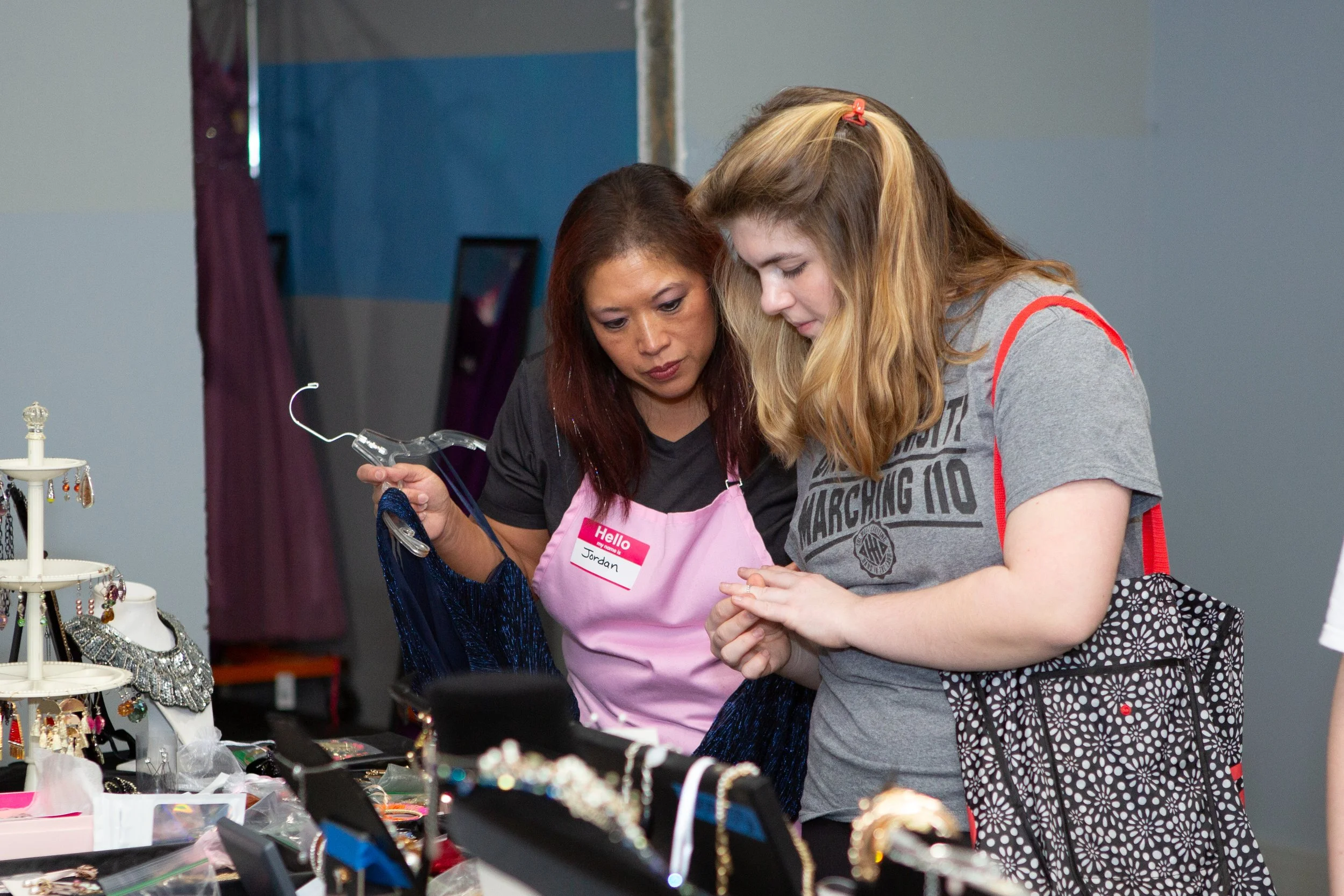 Two women are examining jewelry at a display table, one woman wearing a pink apron labeled 'Hello, my name is Jordan'. The jewelry includes earrings and necklaces, and they are indoors at a craft or jewelry fair.