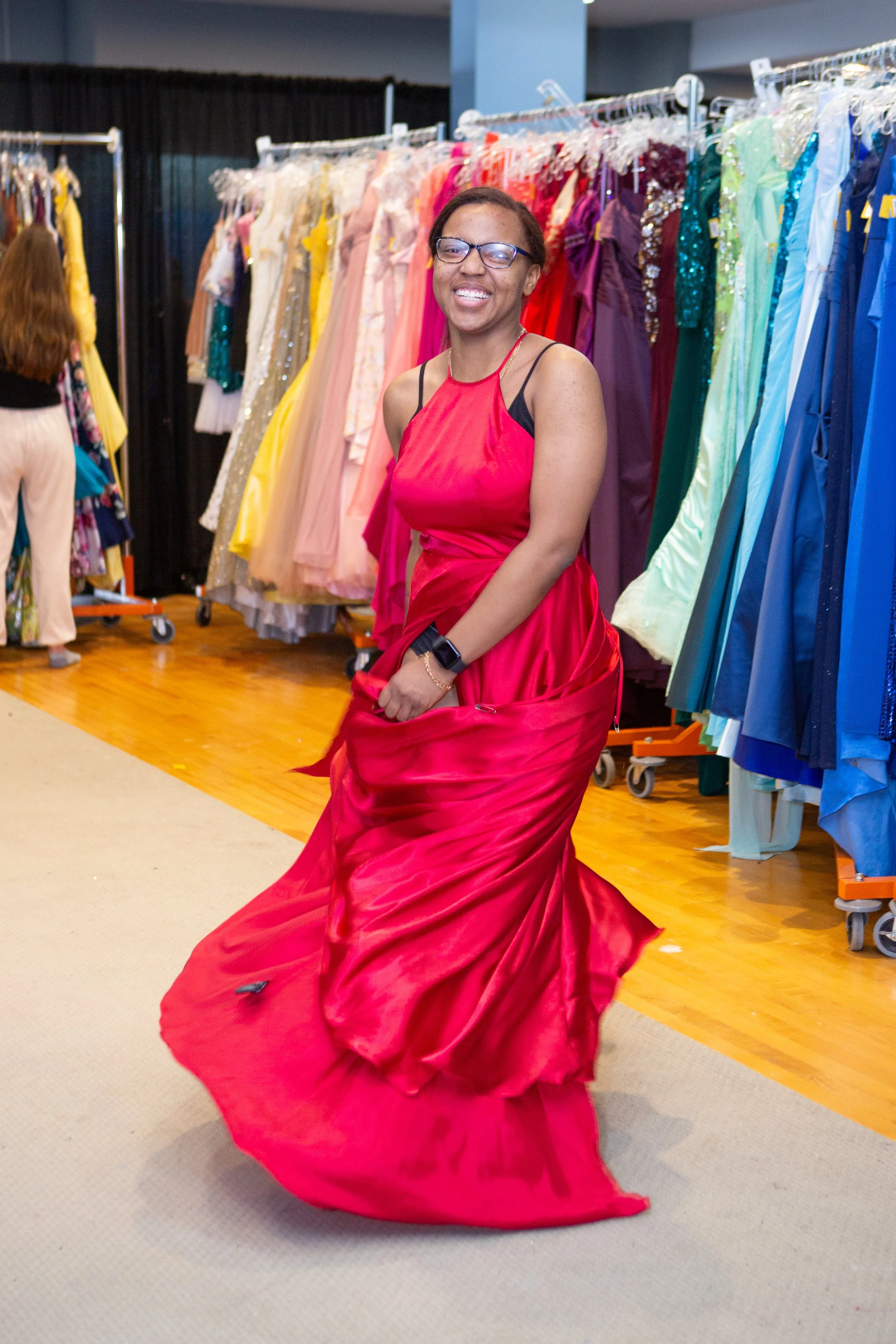 A woman in a red dress, smiling, inside a dress store with colorful dresses hanging on racks in the background.
