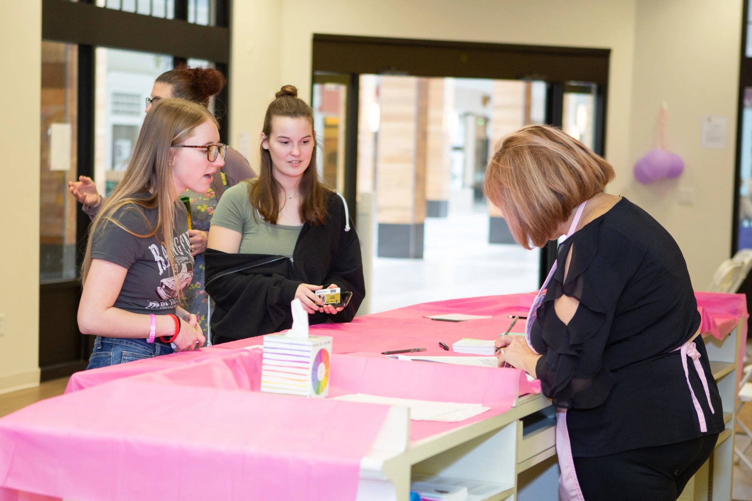 People at a registration or check-in table, with a woman signing in or filling out a form, at an event with pink decorations.