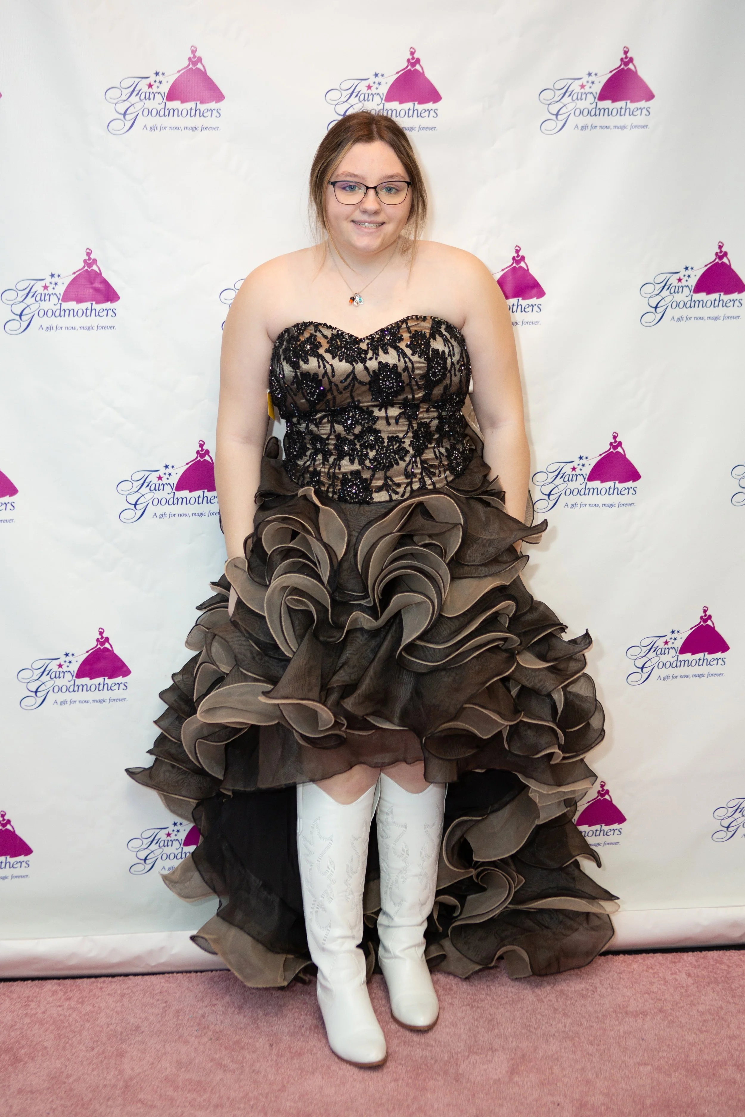 Young woman in a black and beige strapless gown with lots of ruffles, white knee-high boots, and glasses, standing in front of a backdrop with the Fairy Goodmothers logo.
