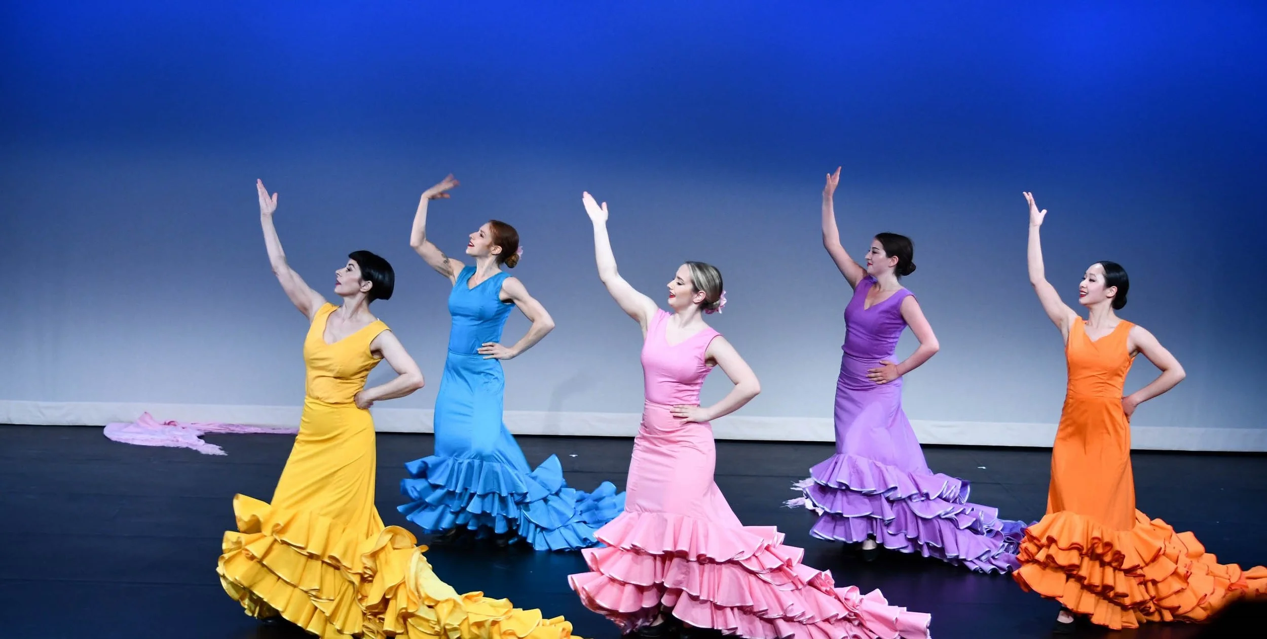Five women in colorful flamenco dresses pose with raised arms on stage against a blue background.