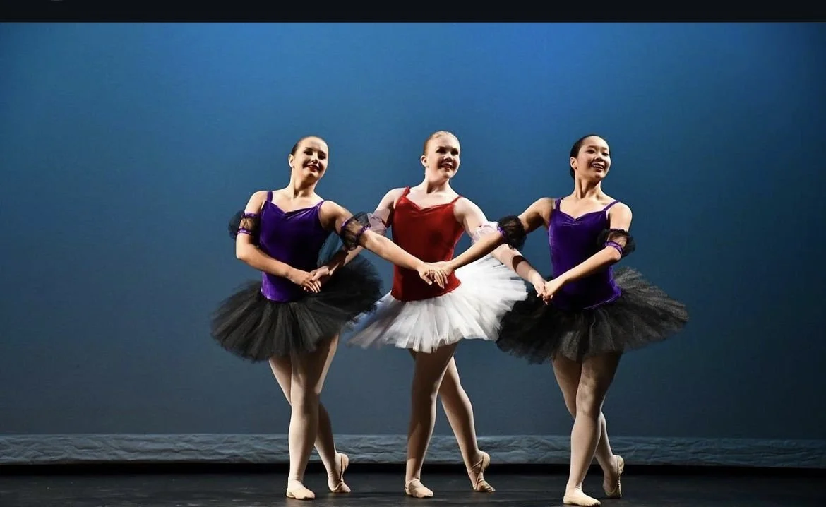 Three ballerinas holding hands, wearing colorful leotards with tutus and ballet slippers, performing on stage against a dark backdrop.