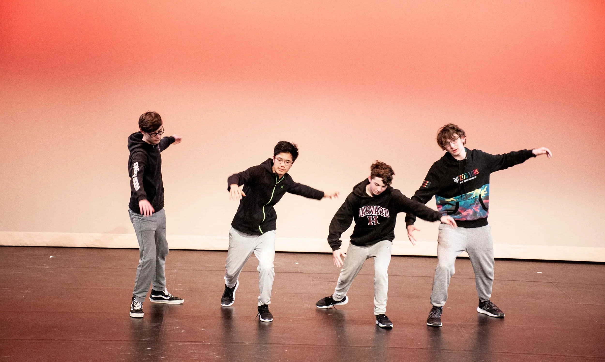 Four young men wearing casual clothes and hoodies are performing a dance on a stage with a plain orange background.