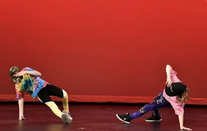 Two children engaged in breakdancing or hip-hop dance moves on a stage with a red background. One child wears a multicolored tie-dye shirt and gold pants, while the other child wears a pink shirt and blue pants with patterns.