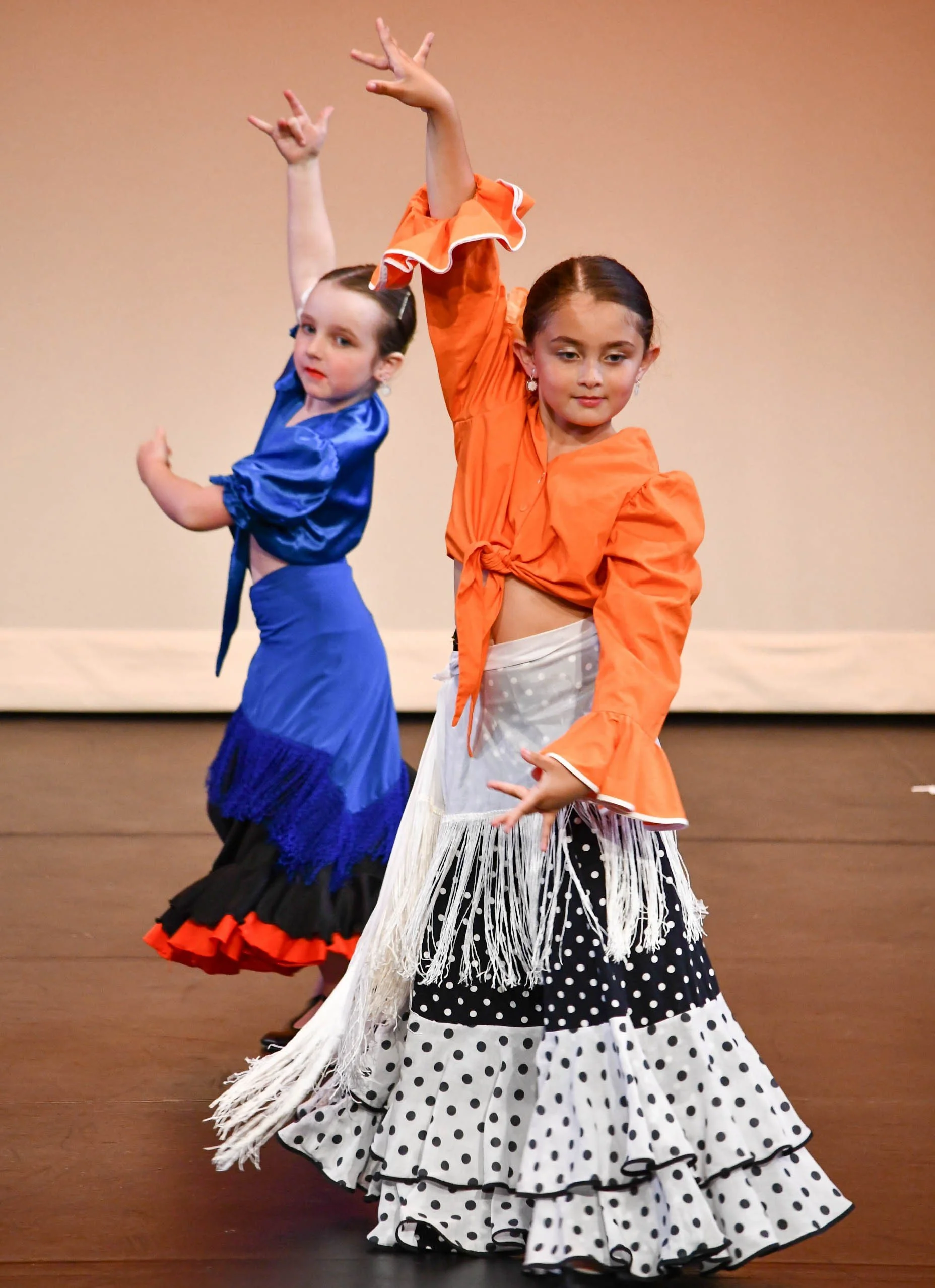 Two young girls performing a dance on stage, wearing colorful dresses with ruffled skirts and blouses, with arms raised in expressive poses.