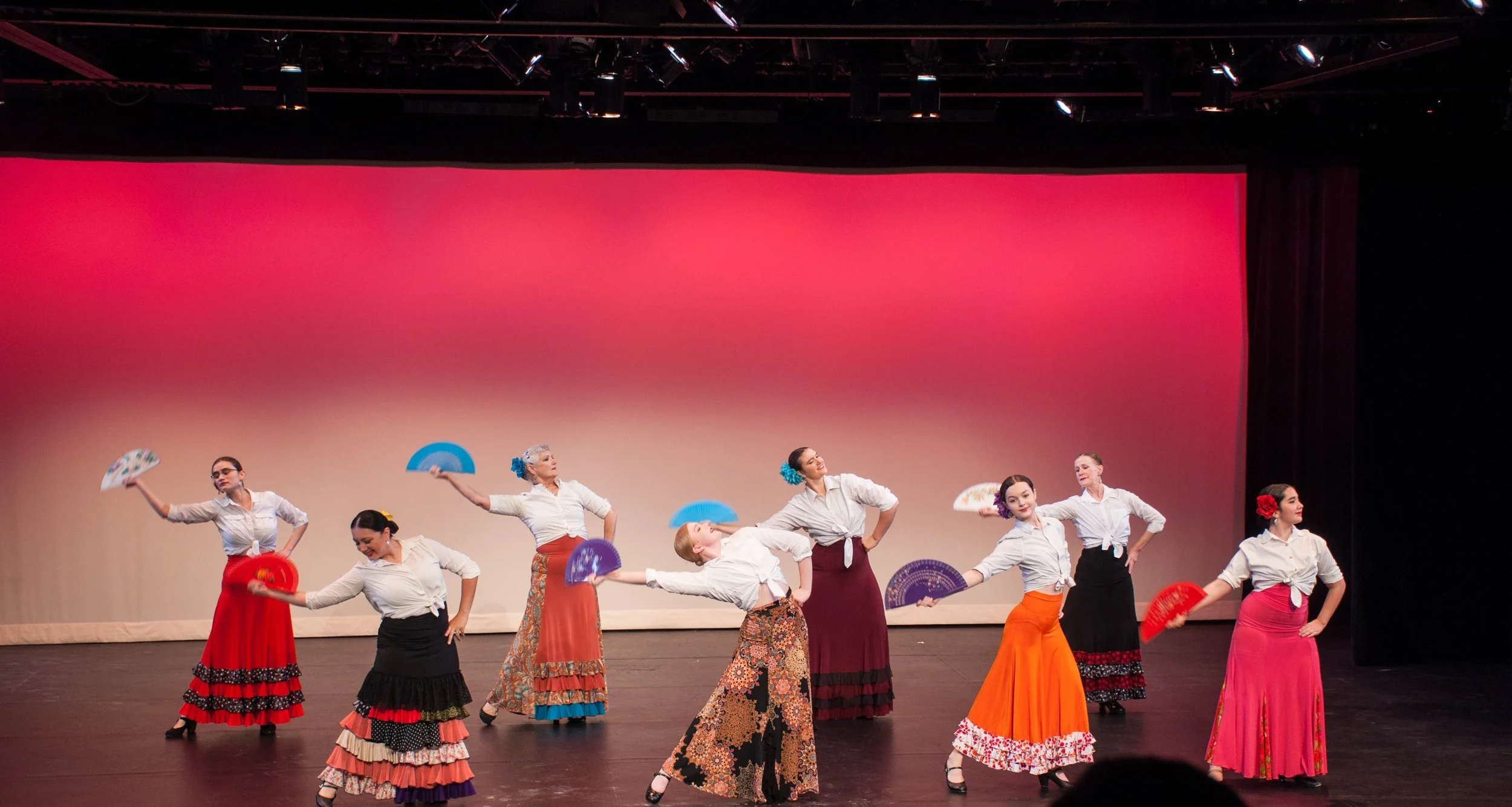 Eight women dressed in traditional Spanish flamenco-style attire performing on stage with colorful fans, with a red and pink gradient backdrop.