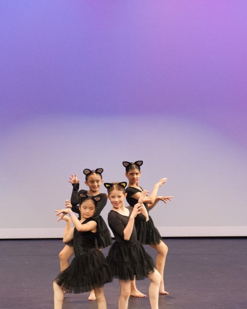 Four young girls dressed in black ballet costumes with cat ear headbands perform a dance on stage.