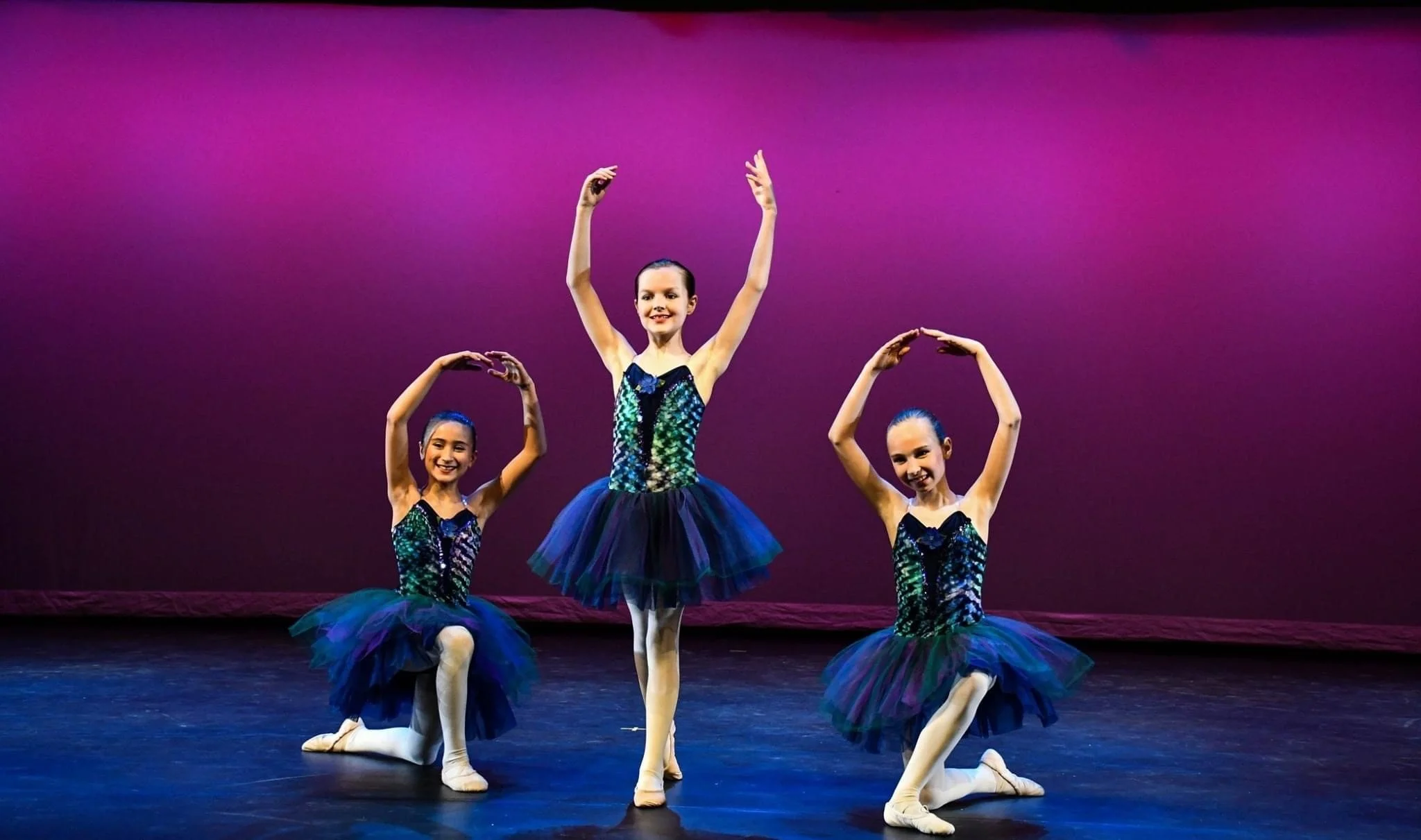 Three young ballerinas in blue tutus performing on stage, with a purple backdrop. Two are kneeling while one is standing, all smiling and holding ballet poses.