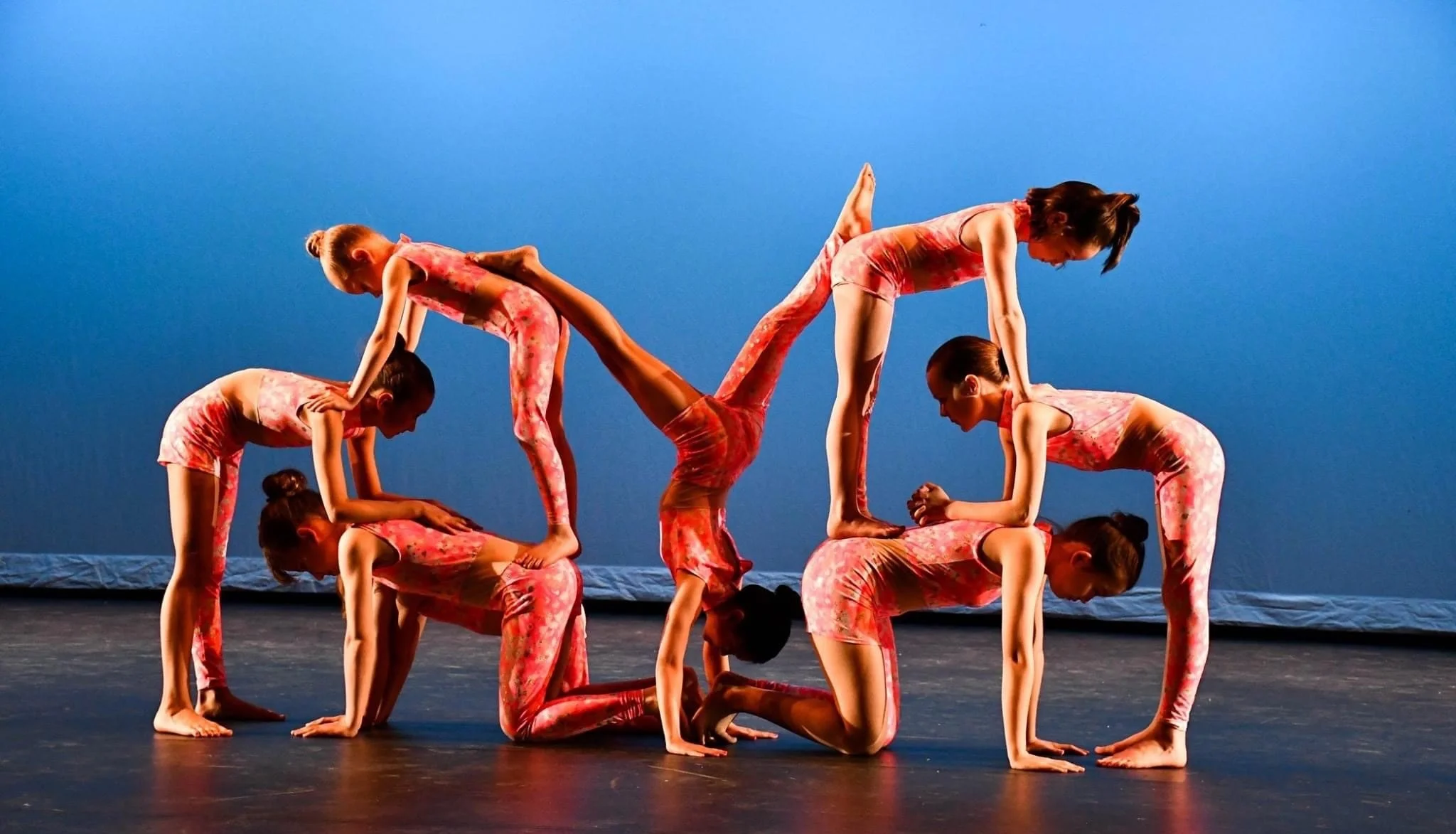 Group of young female gymnasts wearing pink patterned leotards performing coordinated acrobatic poses on a stage with a blue background.