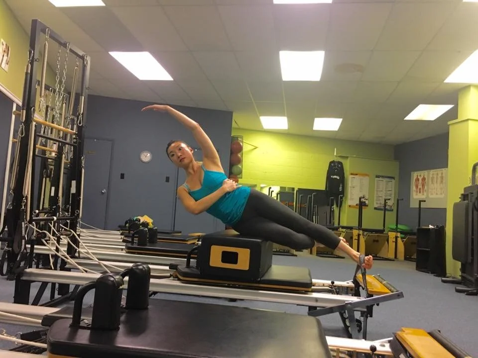 A woman practicing Pilates on a reformer machine in a gym, lying on her side with her upper body supported by her arm and her legs extended, wearing a blue tank top and black leggings.