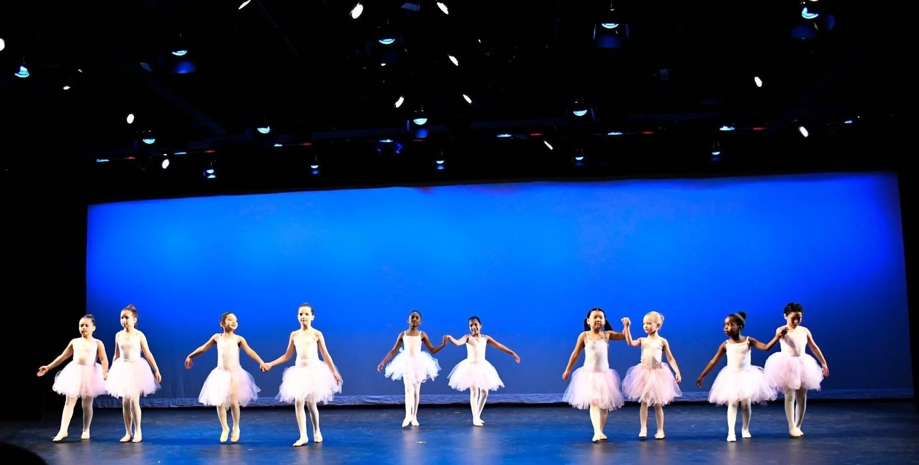 Ten young girls in pink tutus and leotards performing a ballet on stage with a blue background.