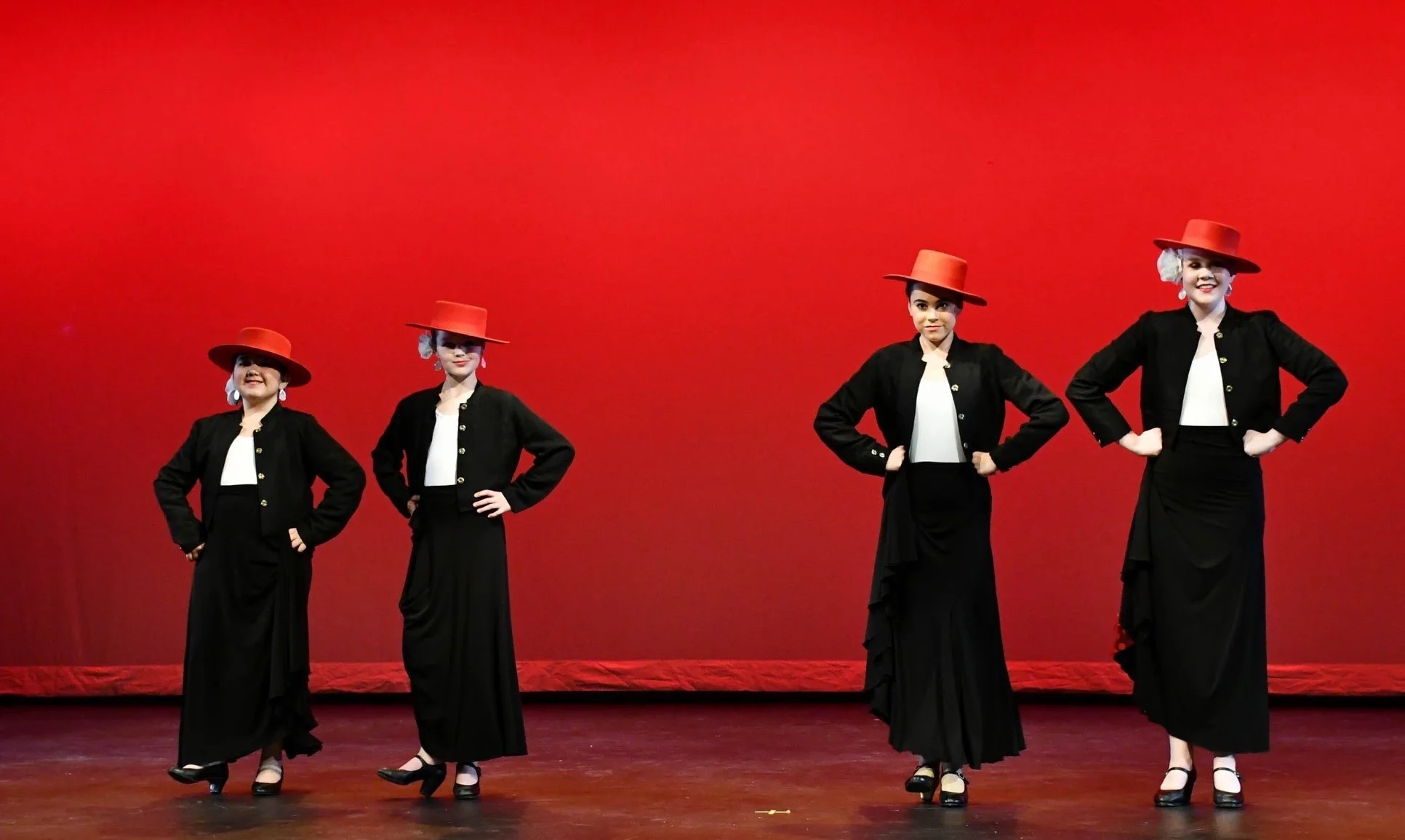 Four women in vintage costumes performing a dance on a stage with a red backdrop, wearing black skirts, black jackets, white tops, red hats, and black heels.