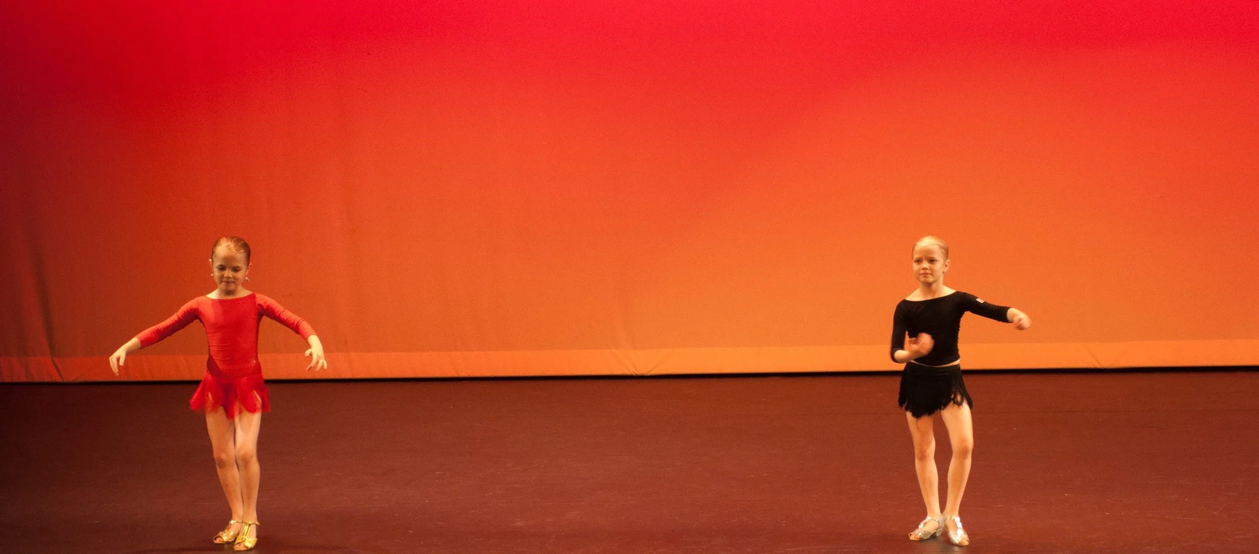 Two young girls in ballet costumes practicing dance on stage with a red-orange background.
