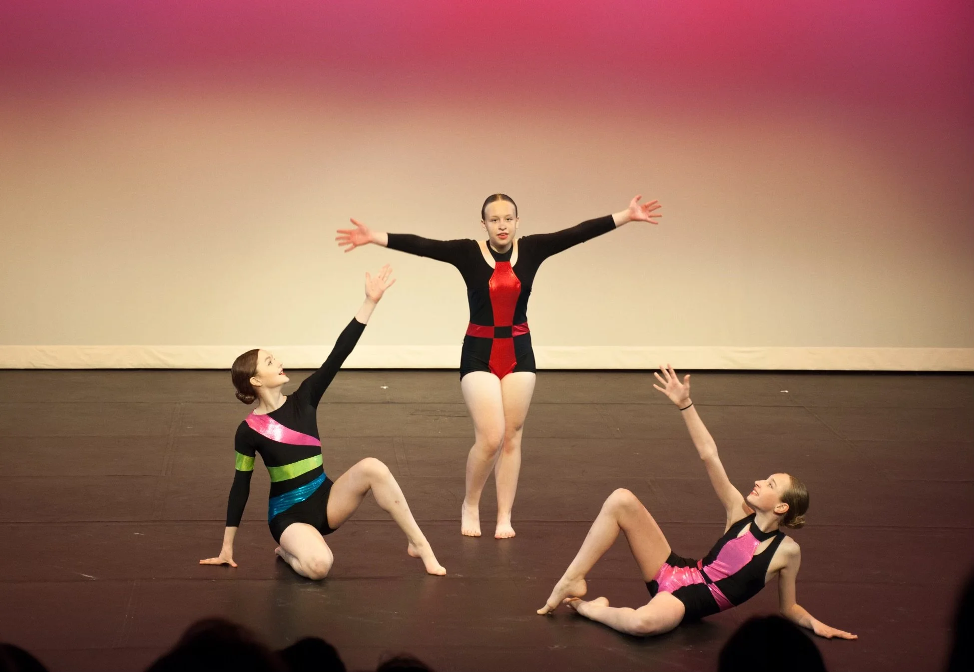 Three young female dancers in colorful costumes perform on stage with a gradient pink and purple background. One dancer stands with arms outstretched, while the other two lie on the floor, reaching toward the standing dancer.