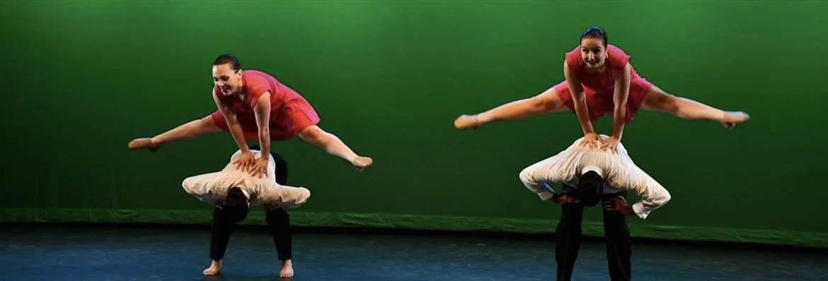 Two performers in red costumes perform an acrobatic dance on a stage with a green backdrop, with each balancing upside down on a person's shoulders.