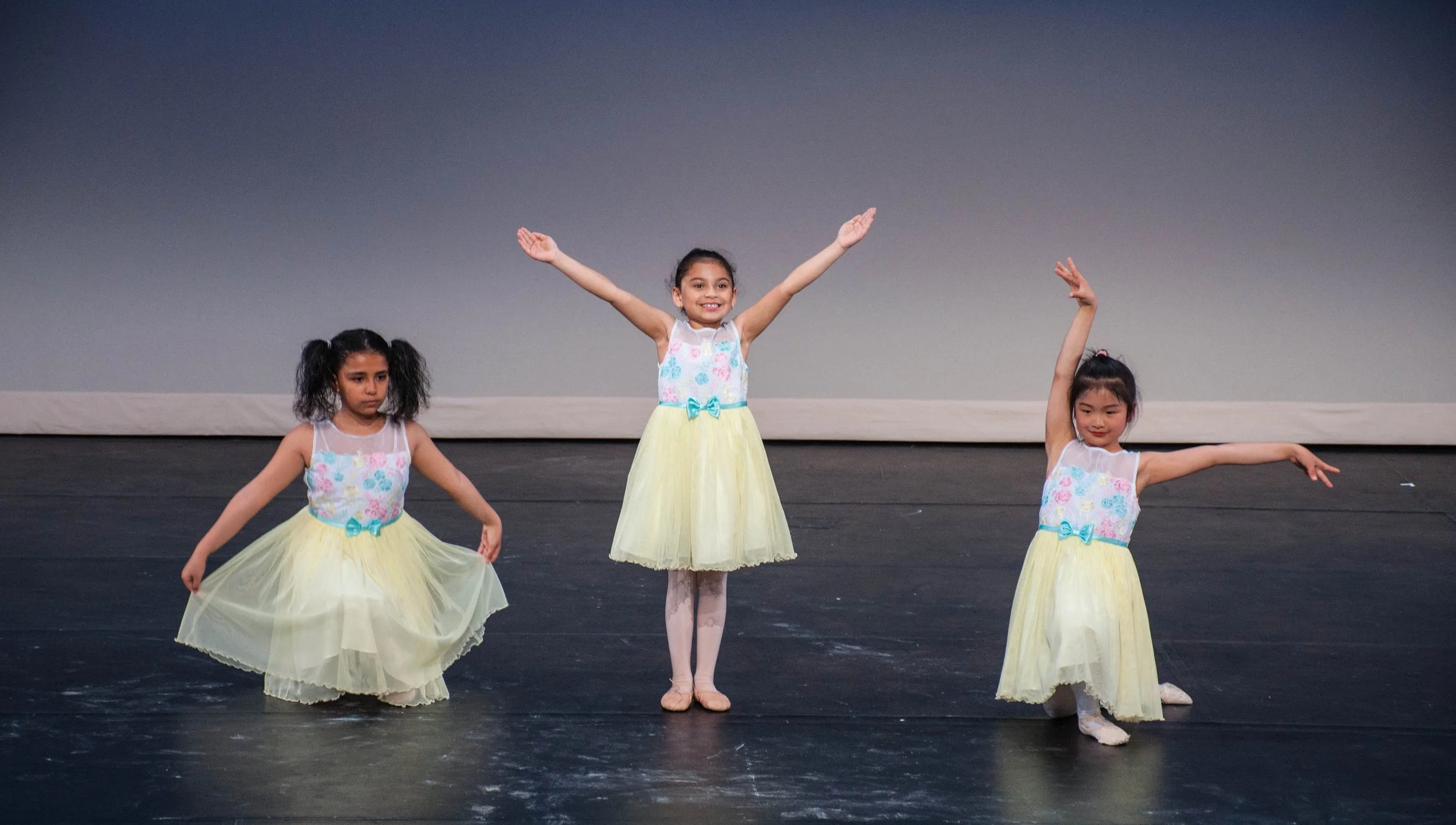 Three young girls in pastel-colored ballet costumes dancing on stage, with one girl in the center raising her arms and smiling, and the other two girl dancers in poses.