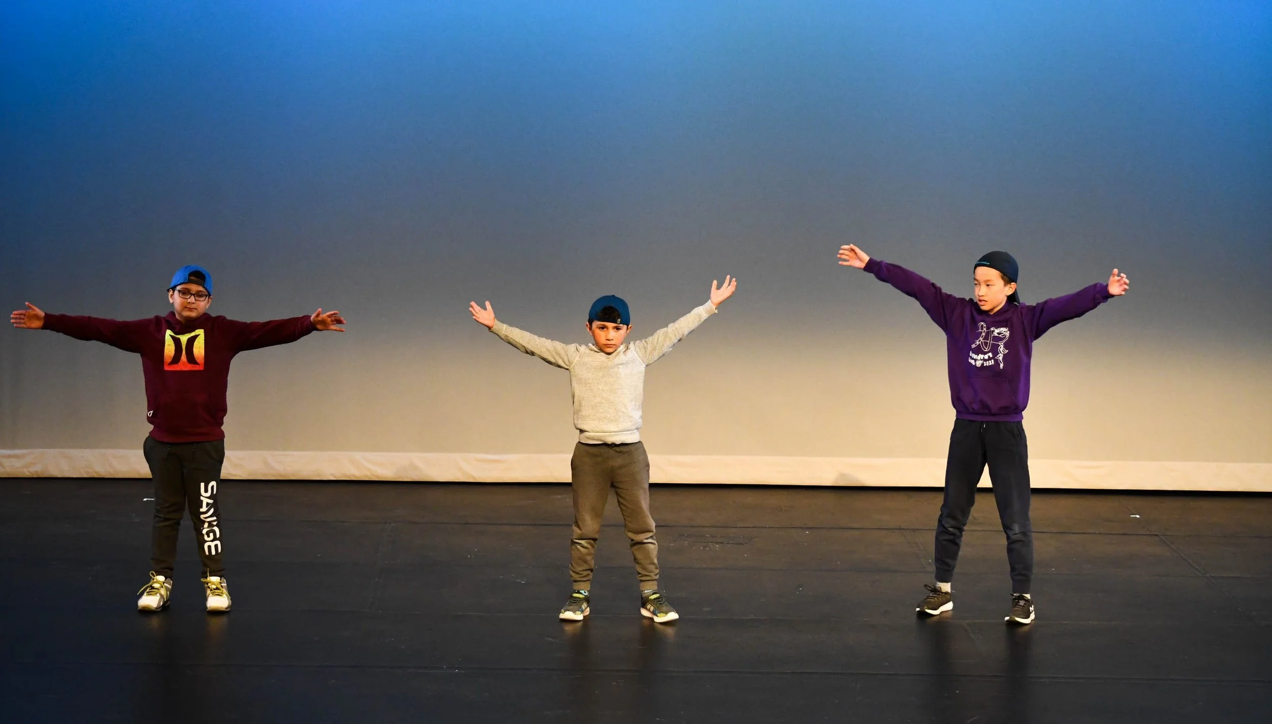 Three boys standing on a stage with arms outstretched, wearing hoodies and caps.