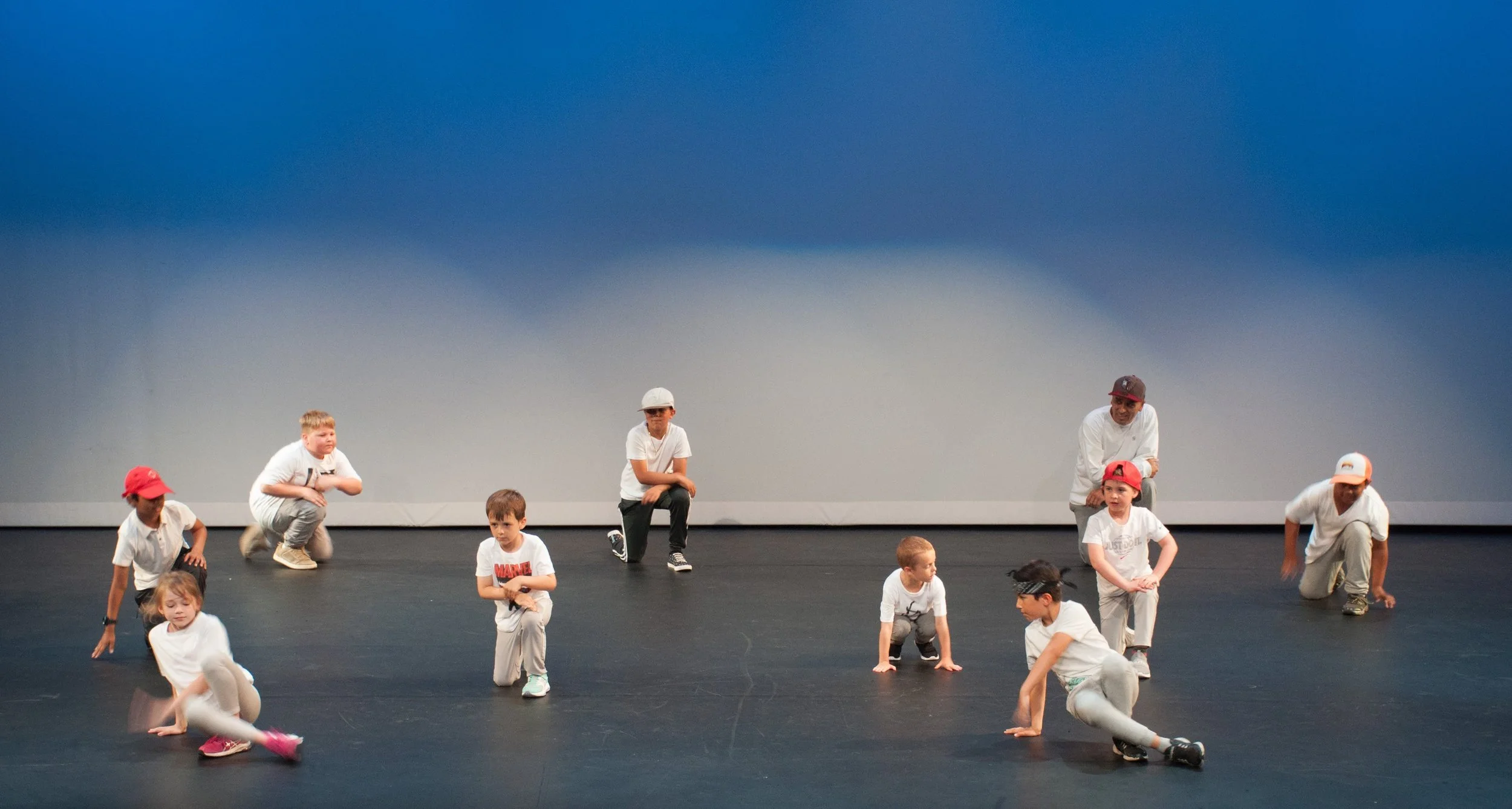 Children and adults performing a dance routine on stage with a plain blue background.