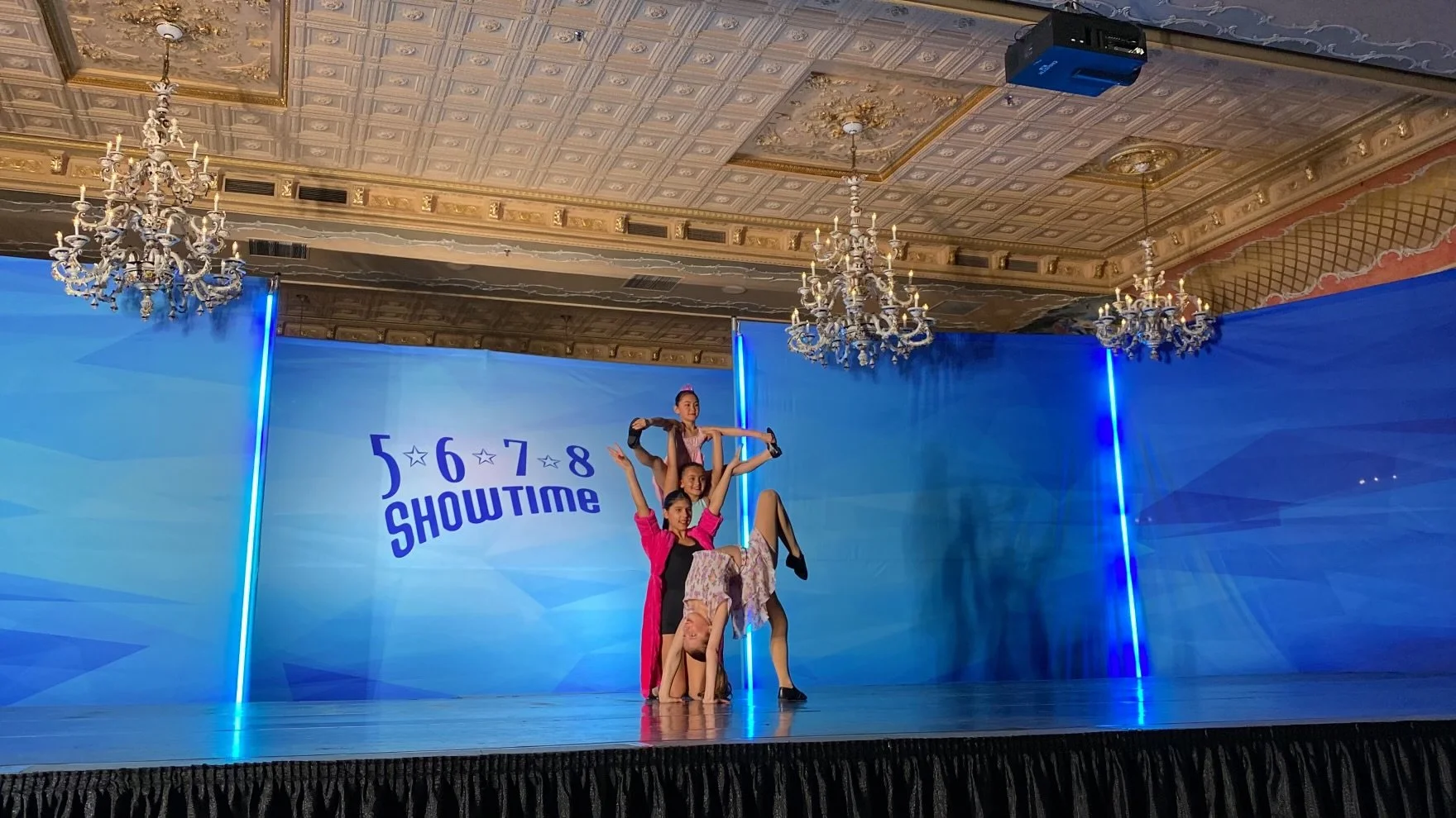 Four female dancers performing a routine on a stage with blue backdrop and chandelier lighting in a ballroom. The backdrop has the text "5 6 7 8 SHOWTIME".