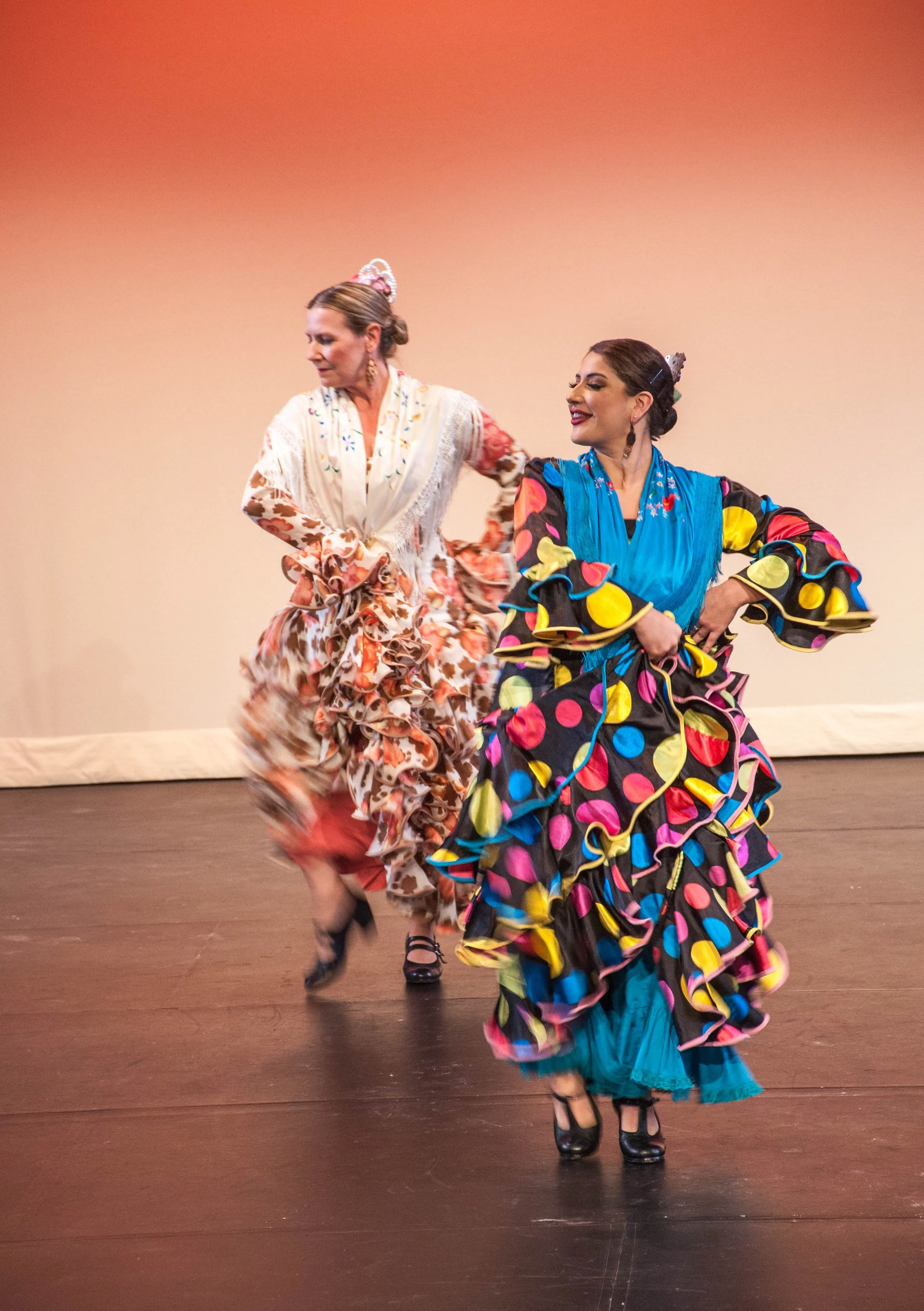Two women dancing in colorful traditional dresses on stage with a plain background.