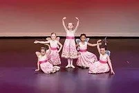 Five young girls in pink and white floral dresses perform a dance on stage, with one standing in the center with arms raised and the others in kneeling or seated poses.