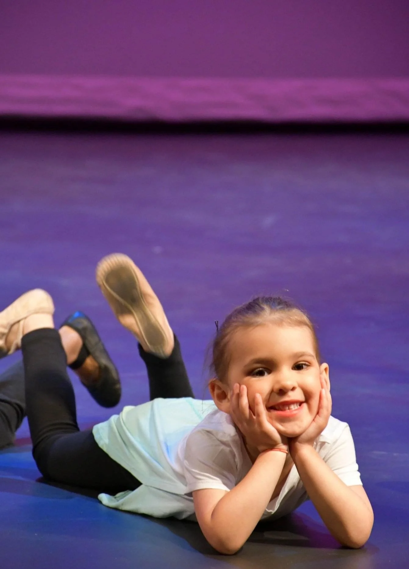 A young girl with a big smile lying on her stomach on a stage floor, resting her head in her hands, wearing a white shirt and black leggings, with her feet raised behind her.