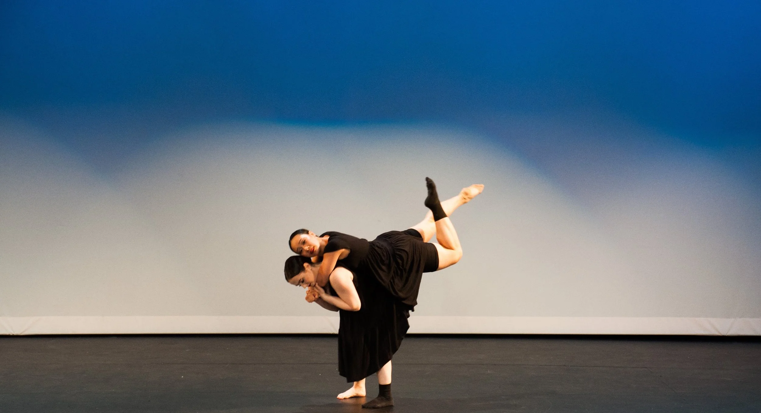 Two dancers performing on stage, one dancer carrying the other on her back, with the stage's gray background.
