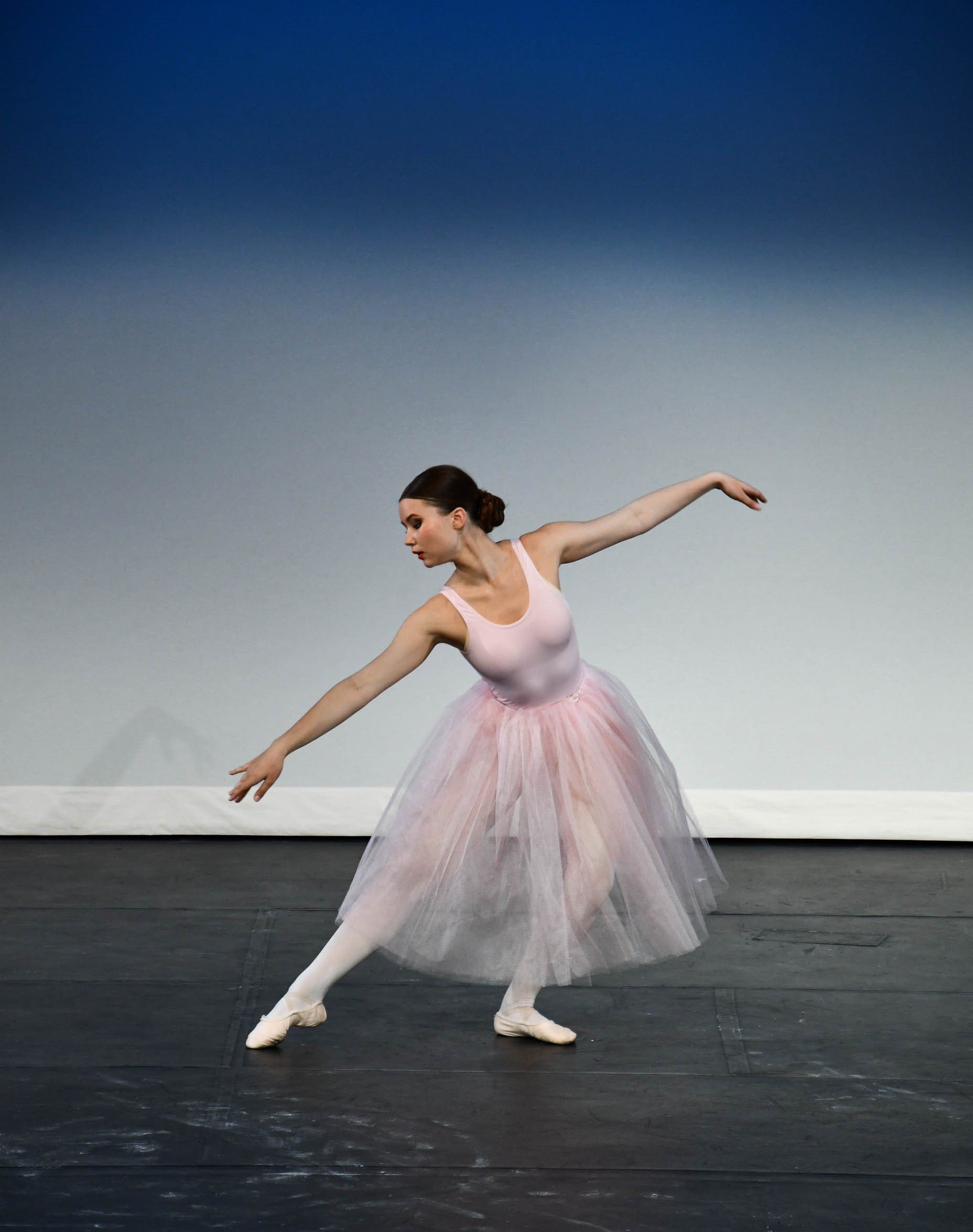 A ballerina in a pink leotard and tutu performing a dance move on stage with a gradient blue background.