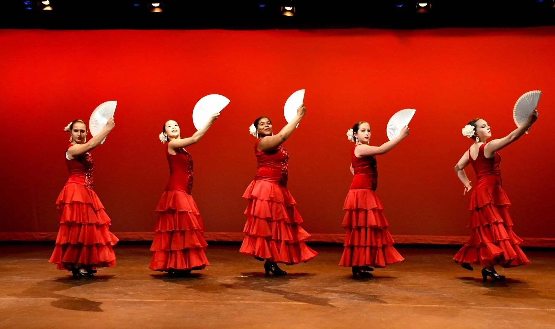 Five women performing a flamenco dance on stage, dressed in red dresses with layered skirts and holding white fans, against a red background.