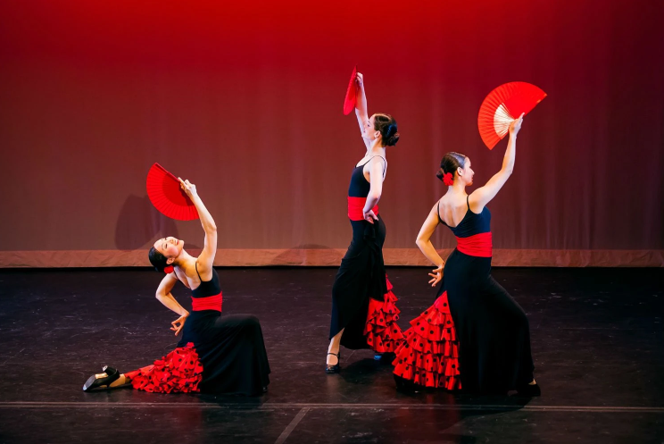 Three women in traditional flamenco dresses performing on stage, each with a red fan, against a red gradient background.