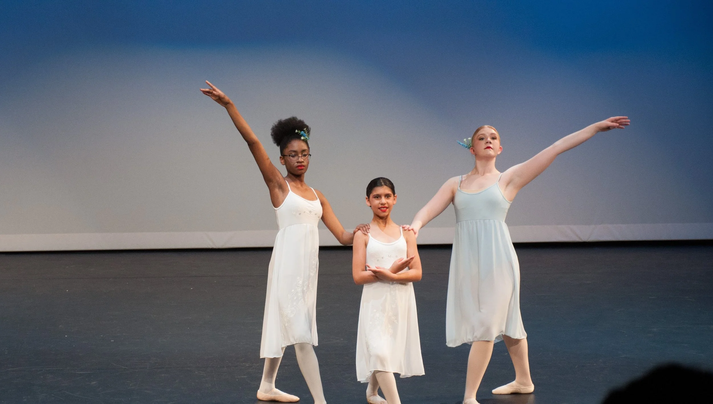 Three young ballet dancers in white costumes on stage, posing with arms extended in a dance formation.