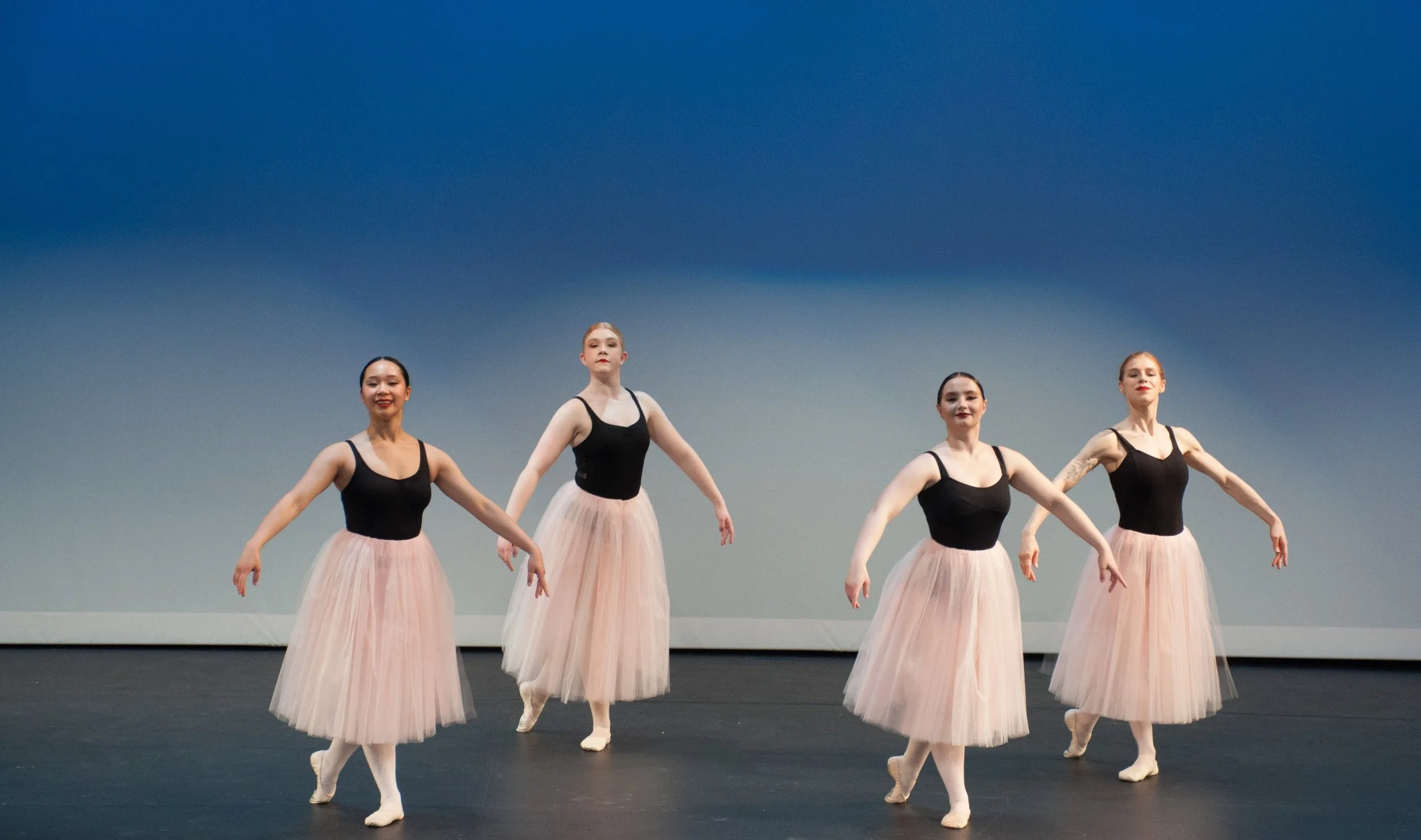 Four ballet dancers in black leotards and pink tutus performing on stage with a blue background.