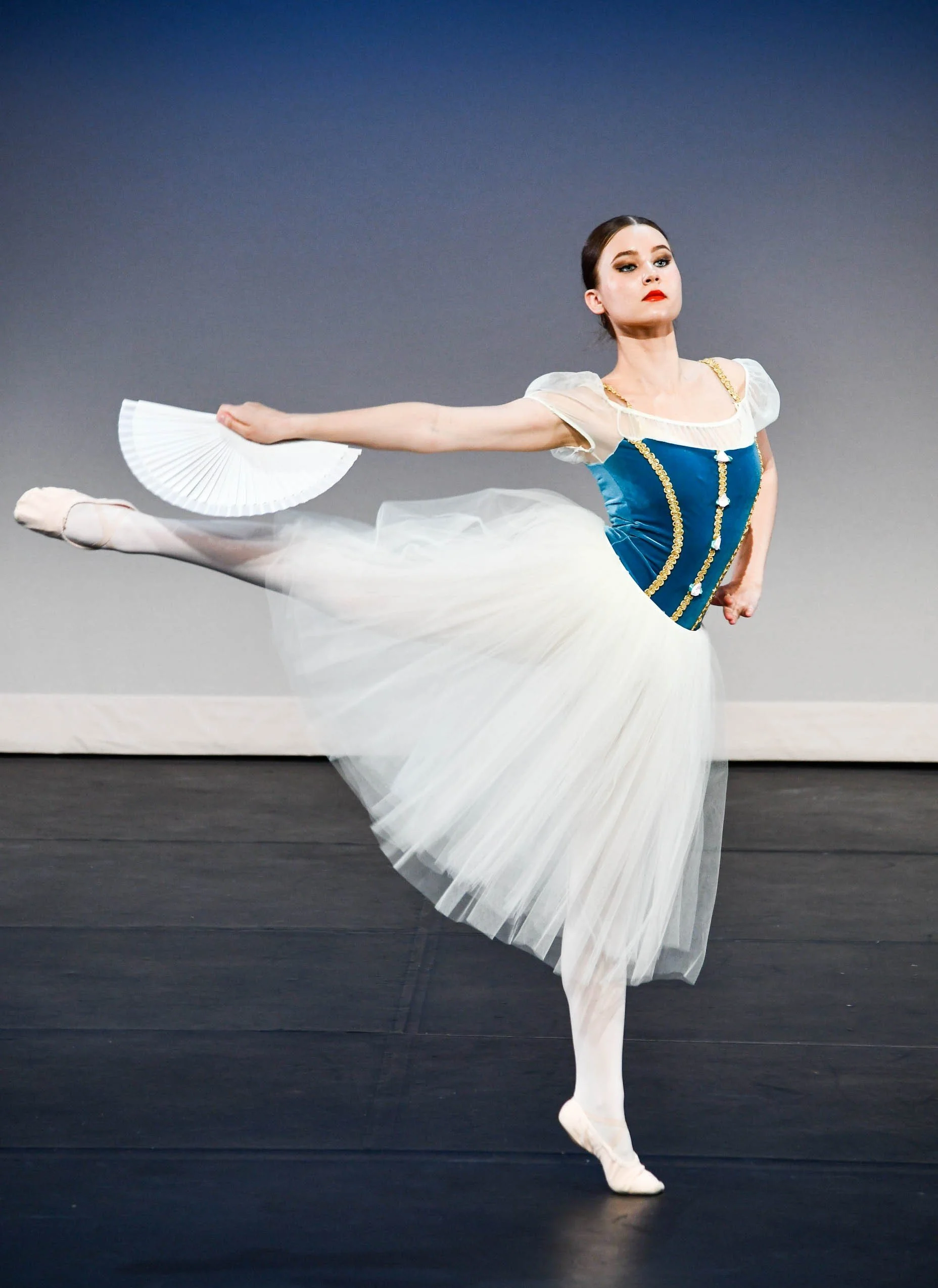 Ballet dancer performing on stage in a tutu and blue bodice, holding a fan, with a neutral background.