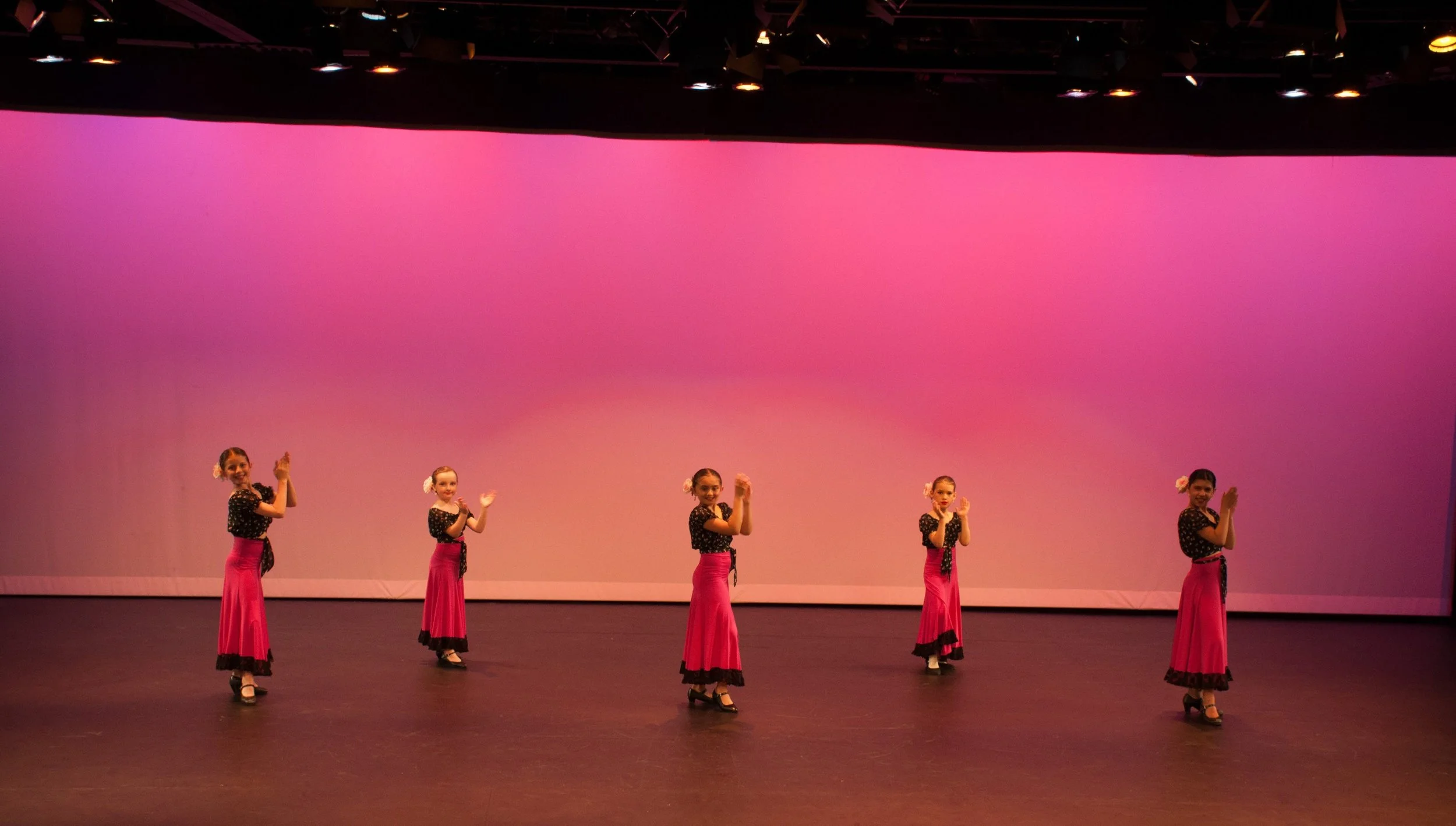 Five young girls dressed in black polka dot tops and pink skirts, performing a dance on a stage with a pink gradient backdrop.