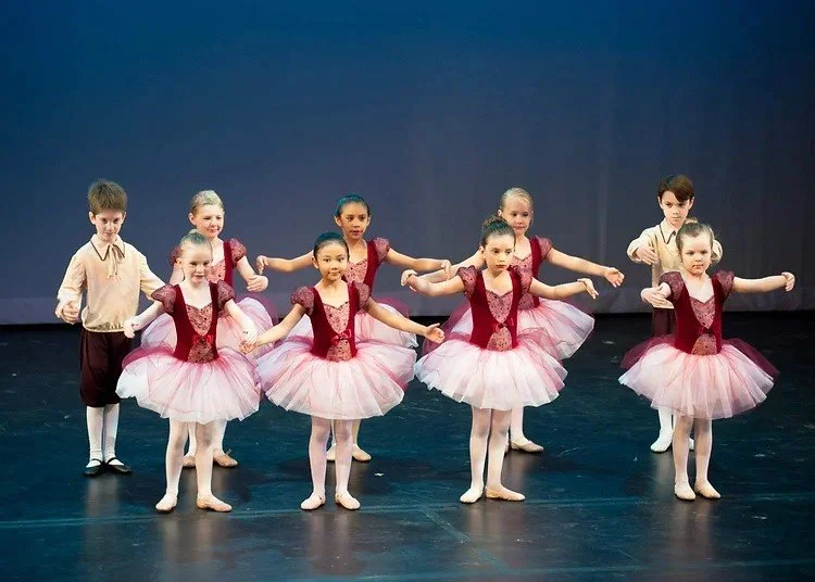 A group of young children performing a ballet dance on stage, wearing pink tutus and costumes.