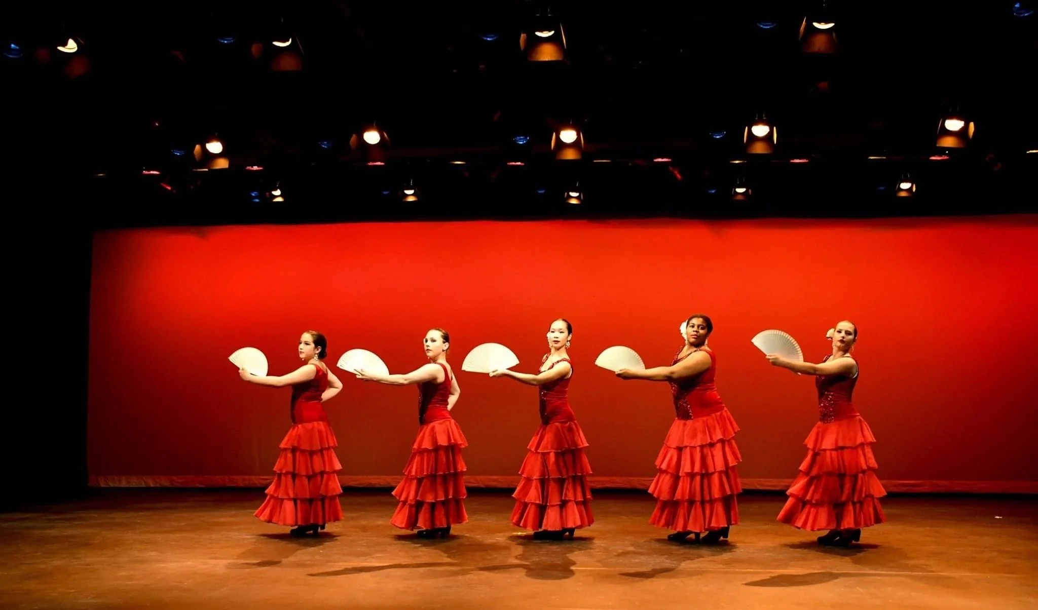Five women in red flamenco dresses performing a dance on stage, holding white fans, against a plain red background.