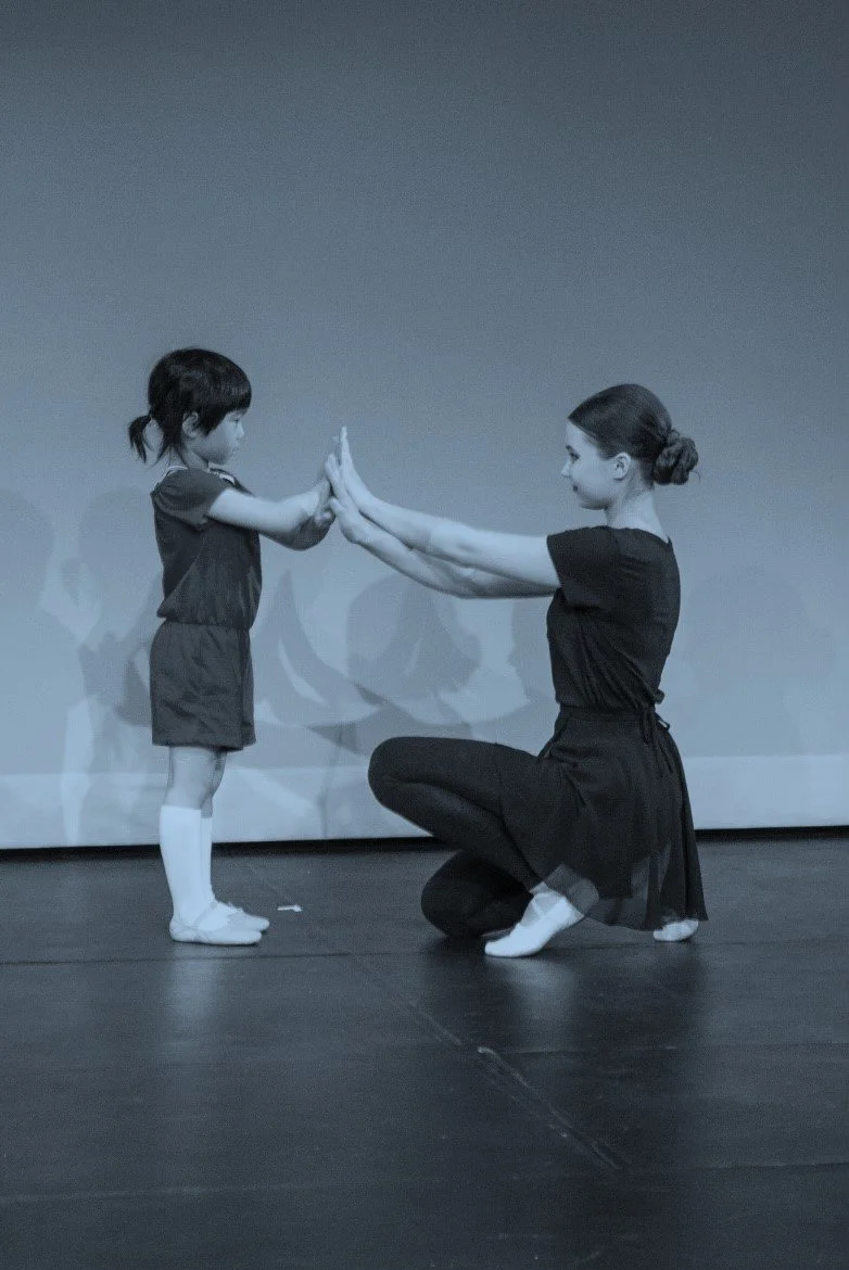 A woman kneeling on one knee, facing a young girl, with both of them touching palms together at face level, in a dance or performance setting.