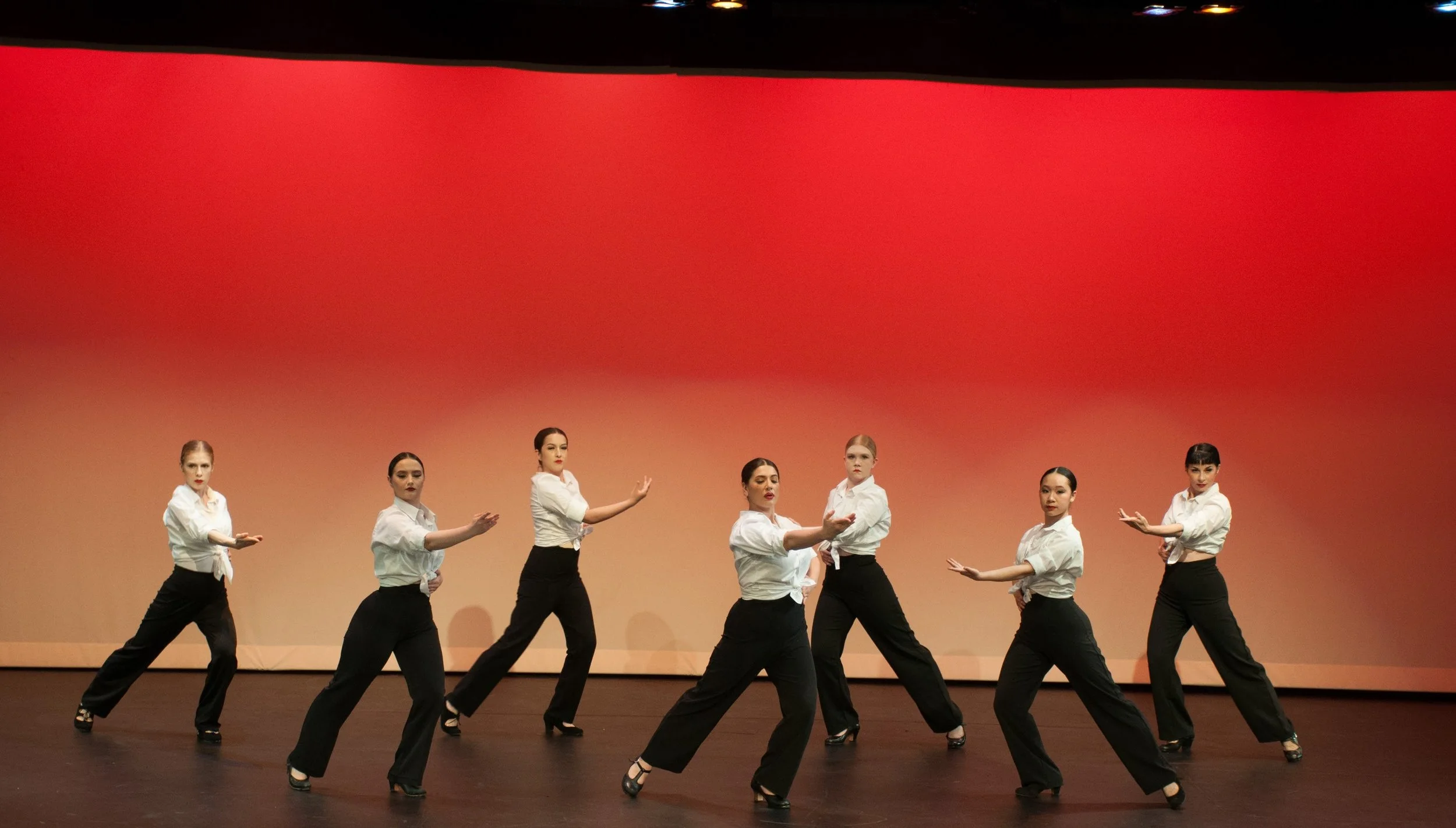 Group of eight women in black pants and white shirts dancing on stage with a red and beige backdrop.