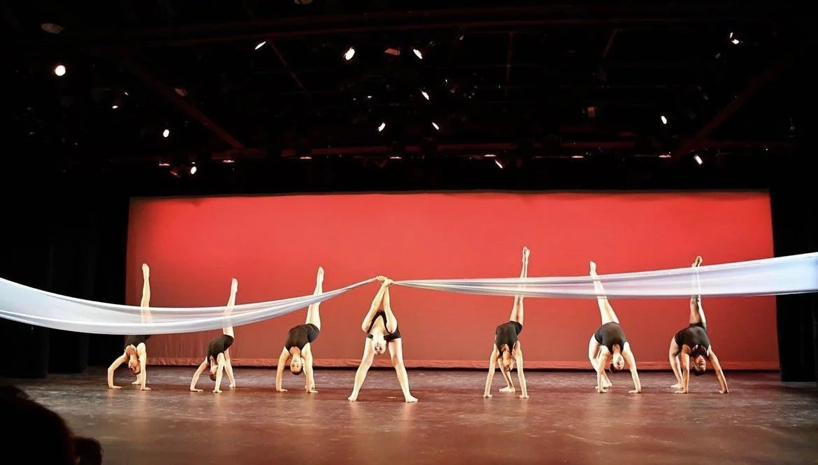 Group of seven dancers performing on stage, some in handstands, holding a long white fabric stretched across the stage, with a red background and black curtains.