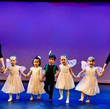 Five children in ballet costumes holding hands on stage, with a purple background.