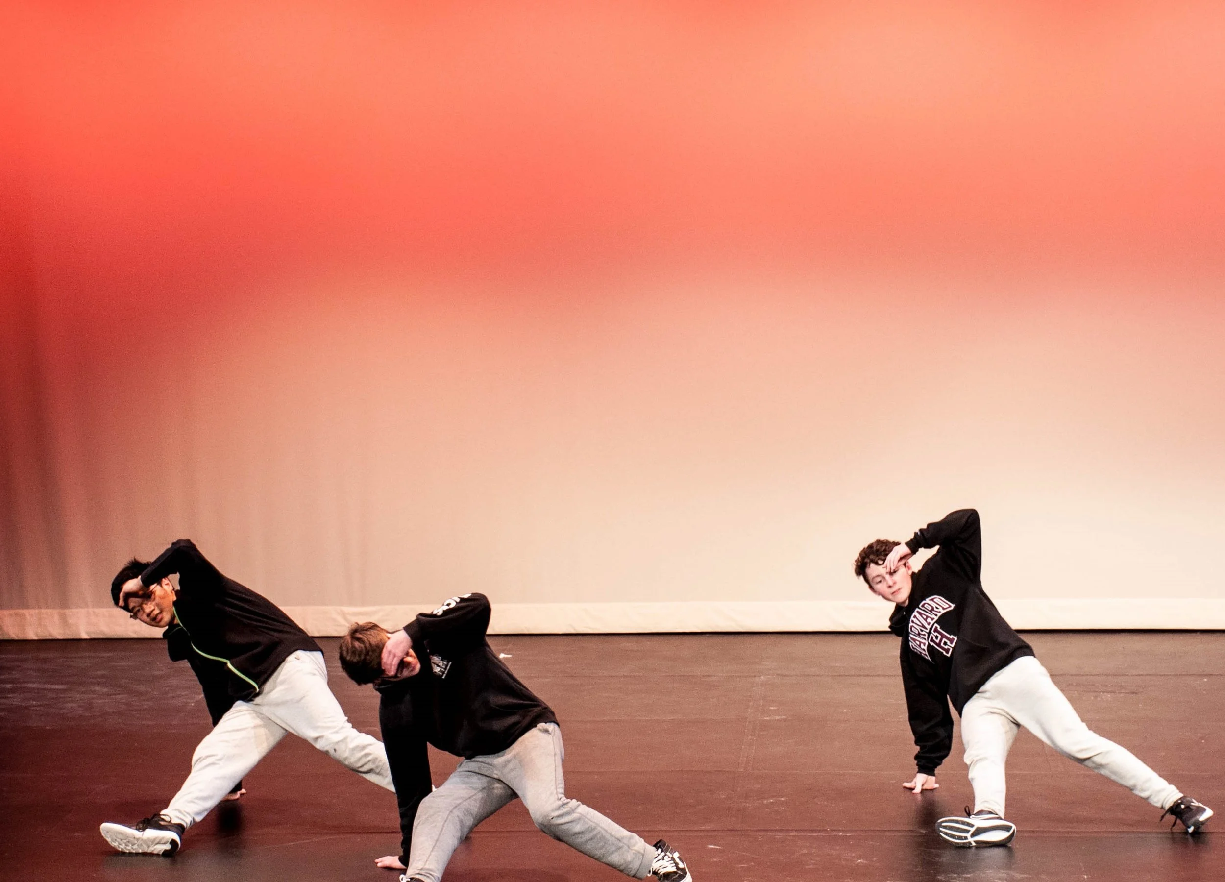 Three young men dancing on stage with a pinkish background, wearing casual streetwear including hoodies and sneakers.