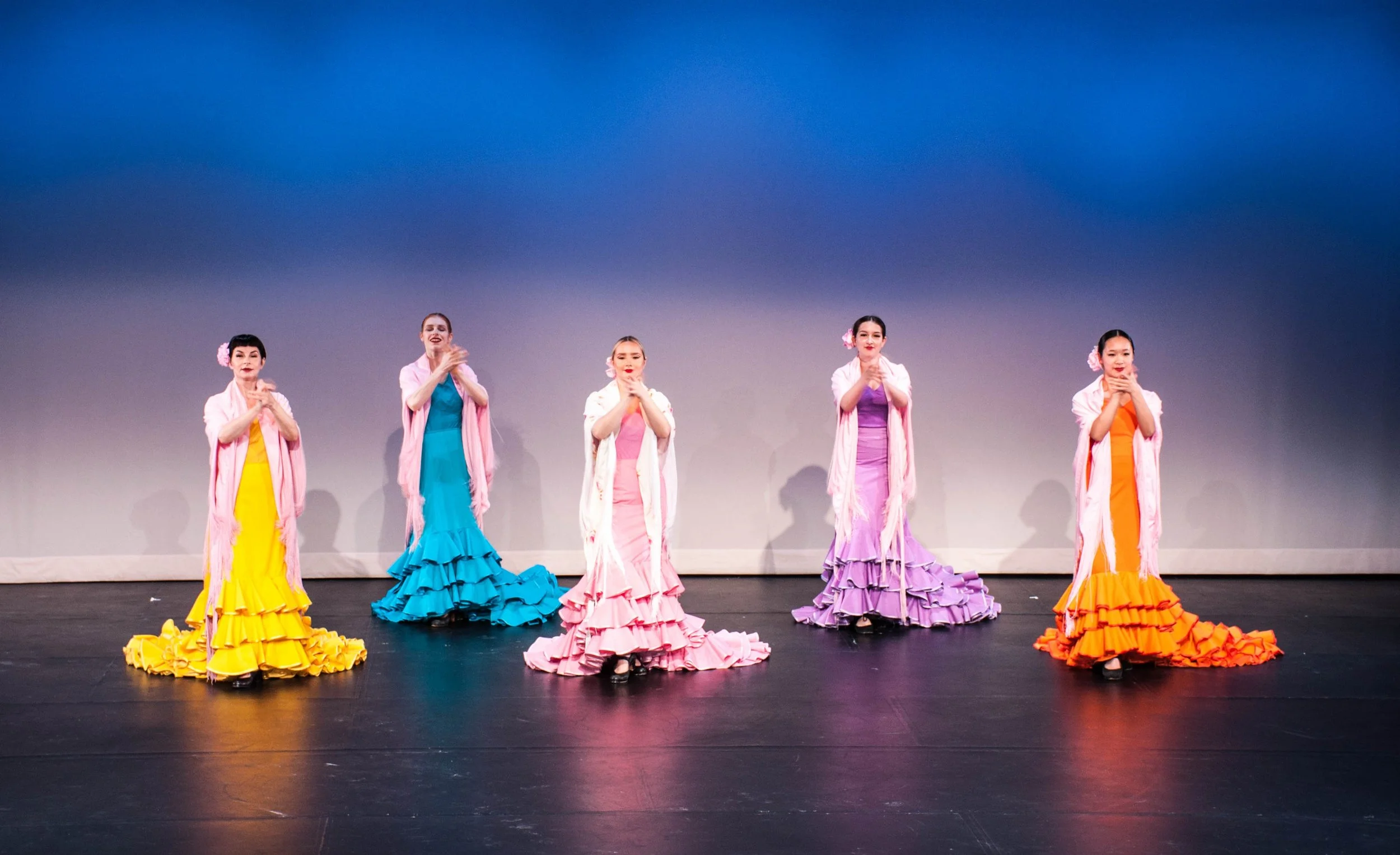 Five women in colorful ruffled dresses performing a dance on stage with a plain gray backdrop.
