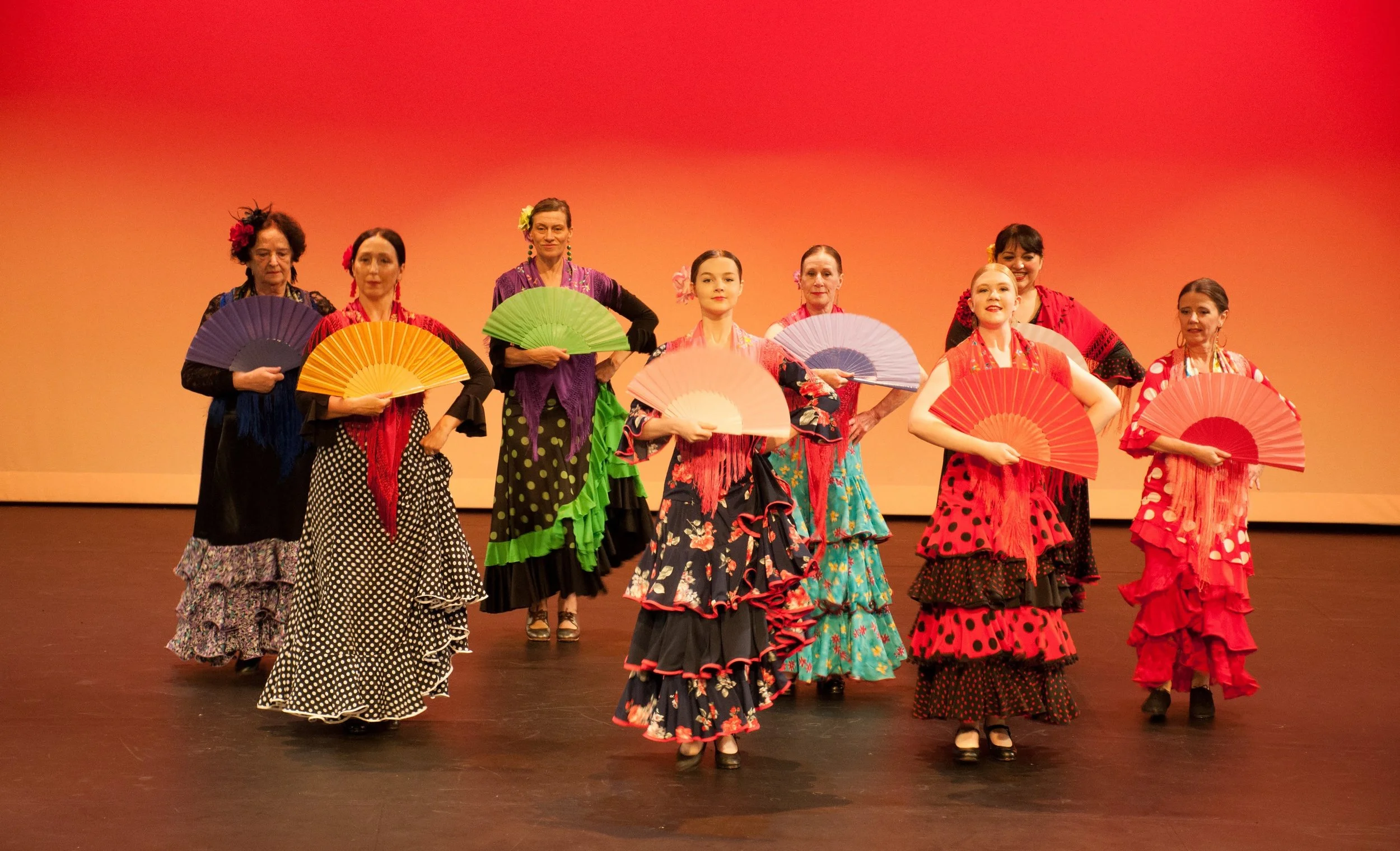 A group of women in colorful flamenco dresses holding fans on stage with an orange background.