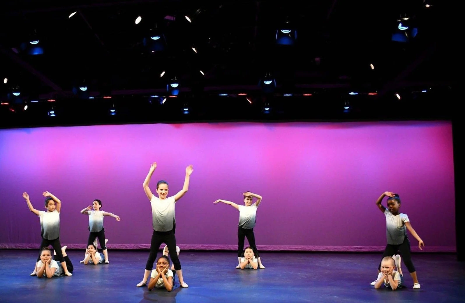 Group of young children performing a dance on stage with a pink background.