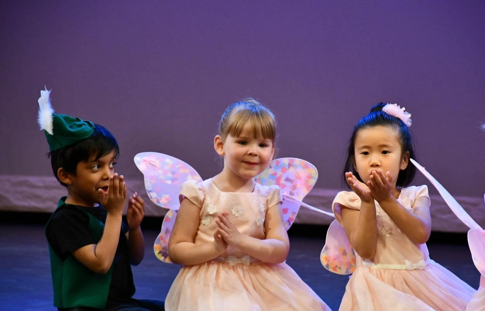 Three young children dressed as fairy tale characters on stage, wearing fairy wings and costumes, with one girl in the center wearing peach-colored dress and fairy wings, flanked by a girl and a boy dressed in costumes, with the girl on the right cov
