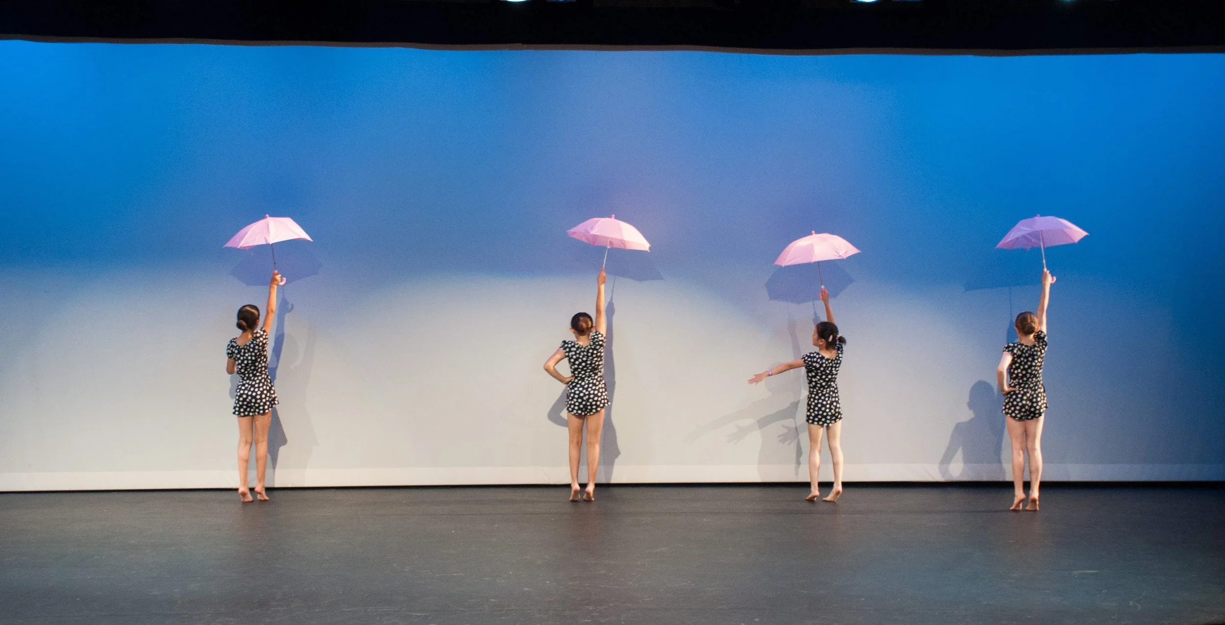 Four women in black and white polka dot outfits holding pink umbrellas during a dance performance on stage with a plain backdrop.