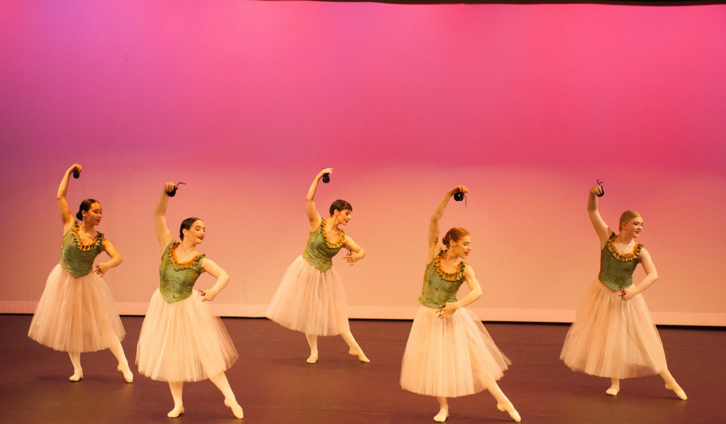 Five ballet dancers in green tops and pink tutus performing on stage against a pink background.