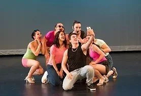 Group of six young performers in colorful costumes taking a selfie backstage.