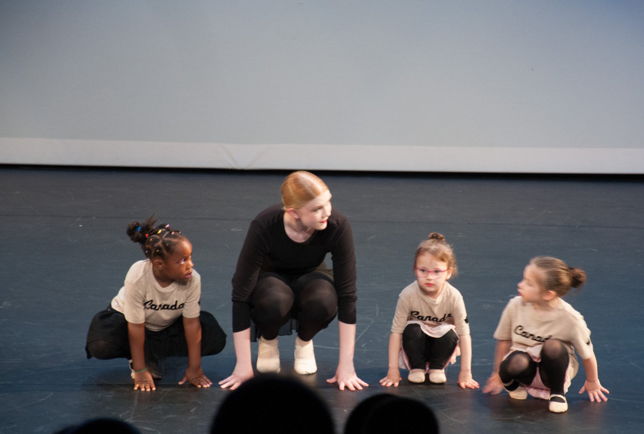 A woman and three young girls crouching on a stage during a performance or rehearsal. The girls are wearing matching beige shirts with 'Canada' written on them, and the woman is in black.