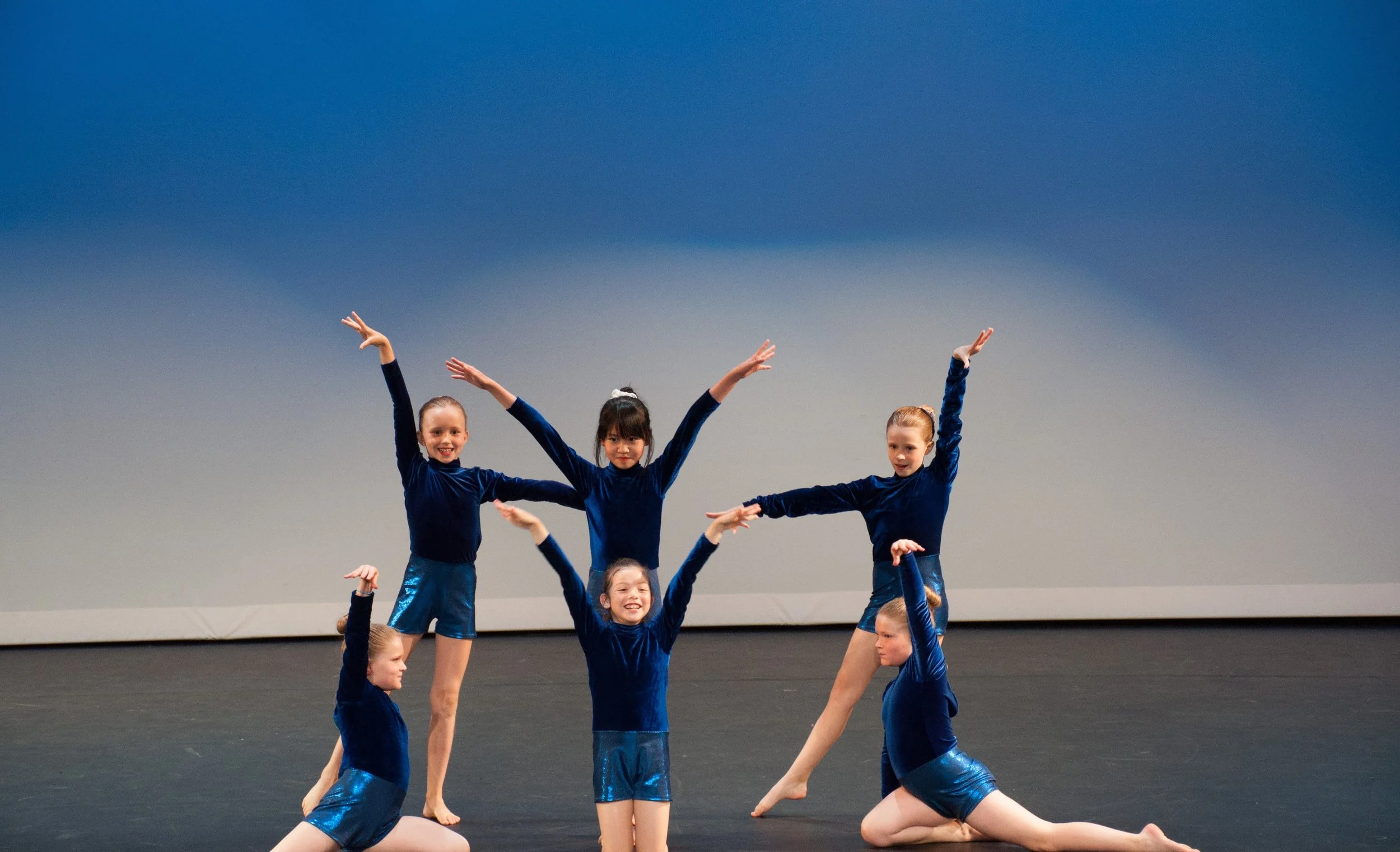 Six young girls in blue dance costumes performing on stage, posing in various ballet positions against a blue background.